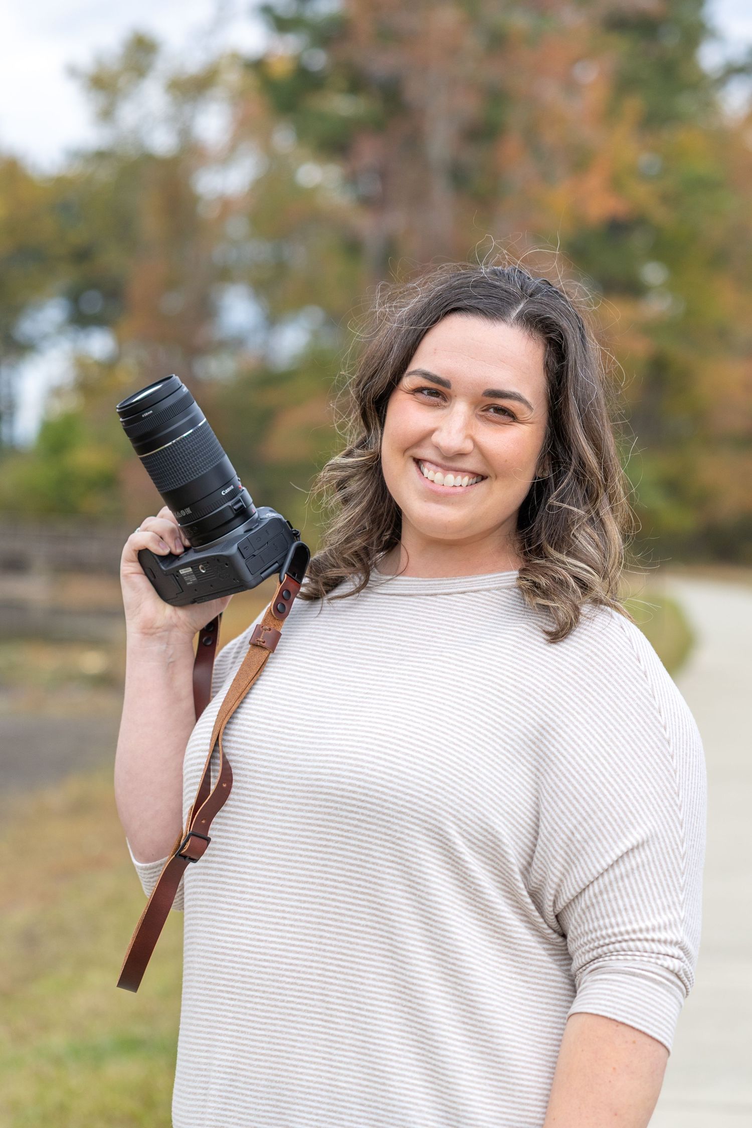 Photographer smiling while holding professional camera with telephoto lens in outdoor autumn setting.
