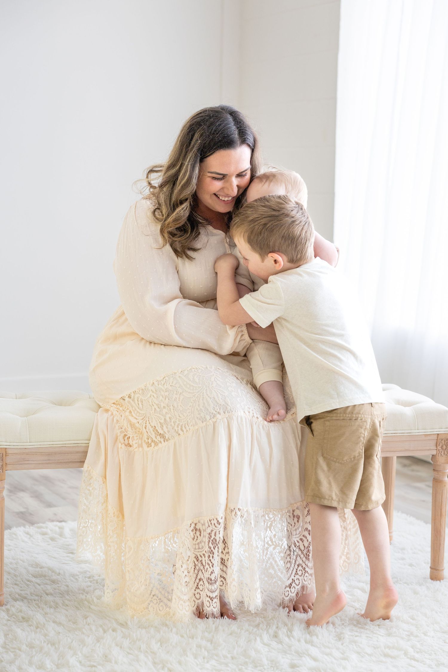 Tender moment captured in bright indoor setting with cream-colored lace dress and natural window light.
