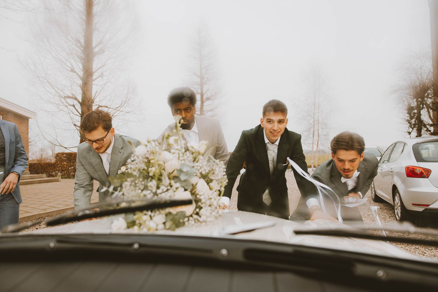 A vintage-style photo shows a group of people in formal attire leaning over a car hood in a playful pose.