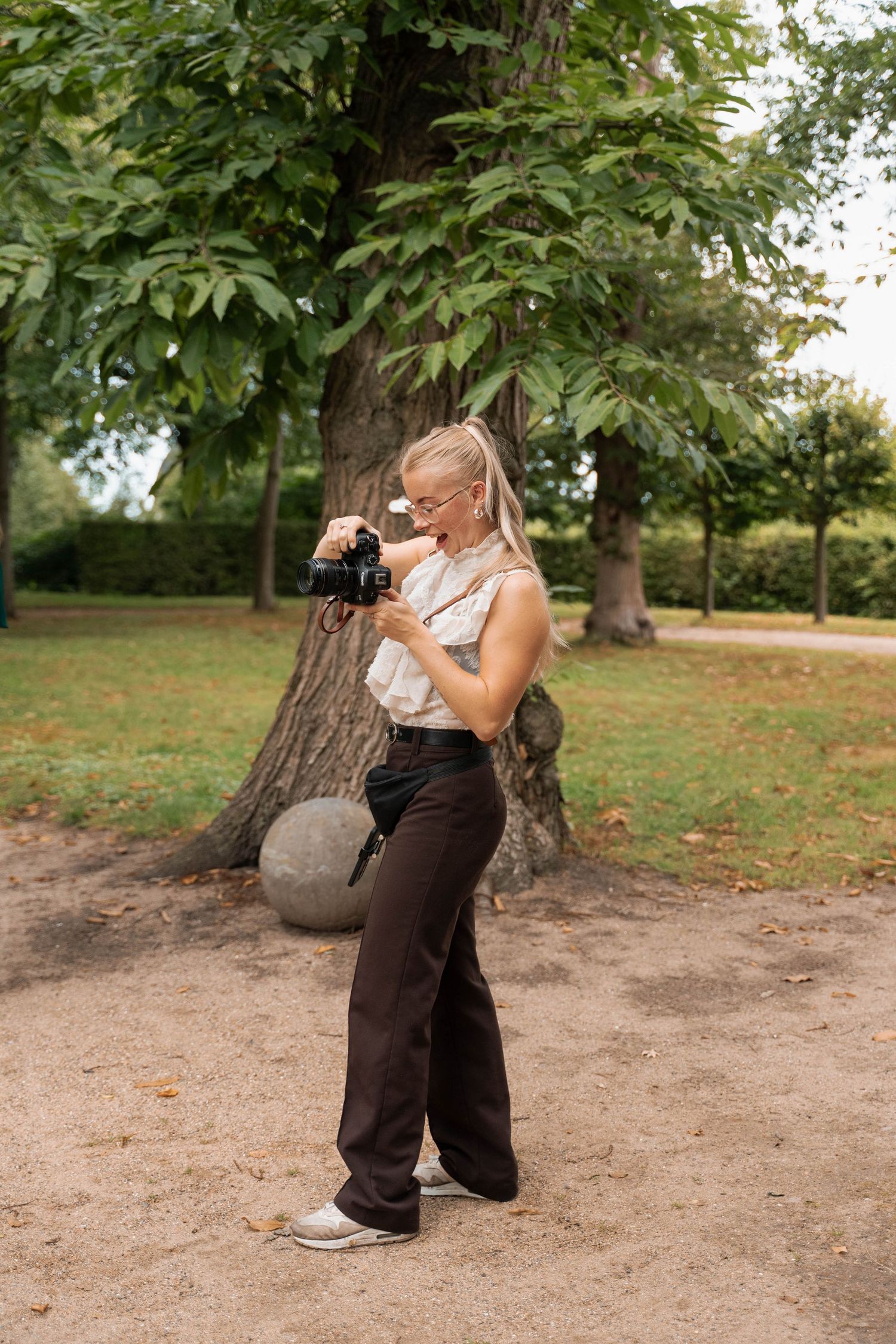 Blonde woman in dark wide-leg pants reviewing photos on camera in a lush green park setting.
