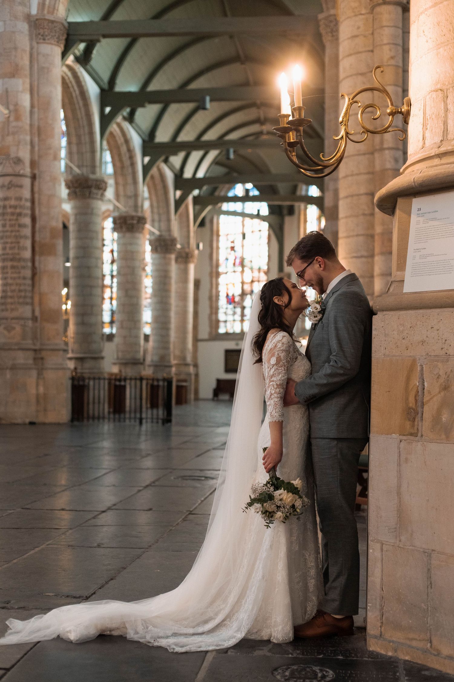 Bride and groom share a romantic moment in a grand cathedral, bride in lace gown with veil, groom in gray suit.