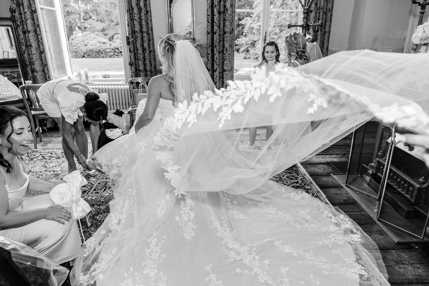 Black and white photo of bridesmaids helping bride with flowing lace wedding dress and veil in elegant room.