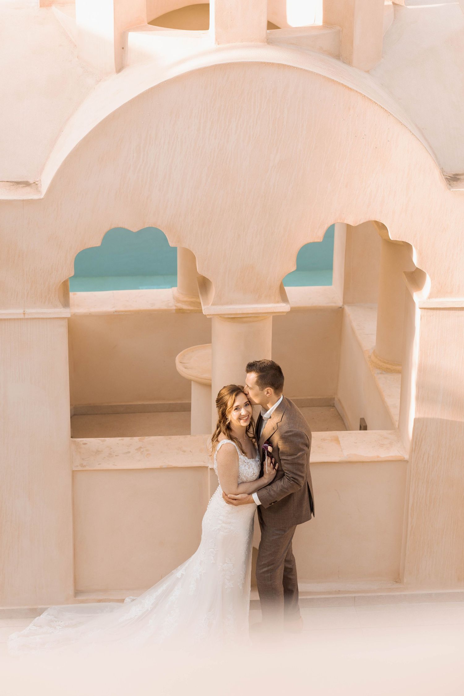 Bride in a lace wedding gown and groom in a brown suit embrace in front of a sunlit peach-colored building with arched turquoise windows.