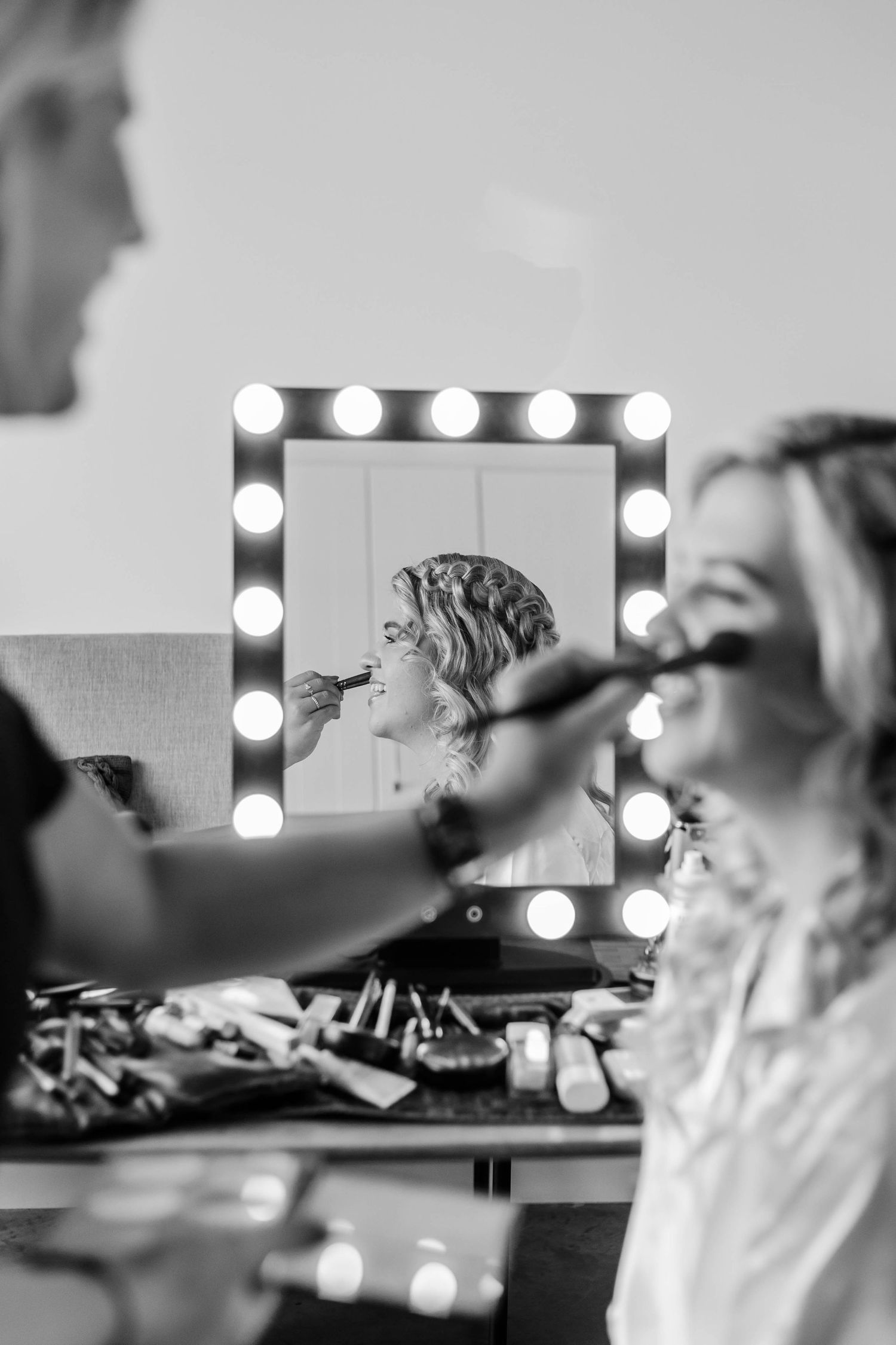 Black and white photo of a makeup artist applying makeup to a smiling bride in front of a lit vanity mirror.