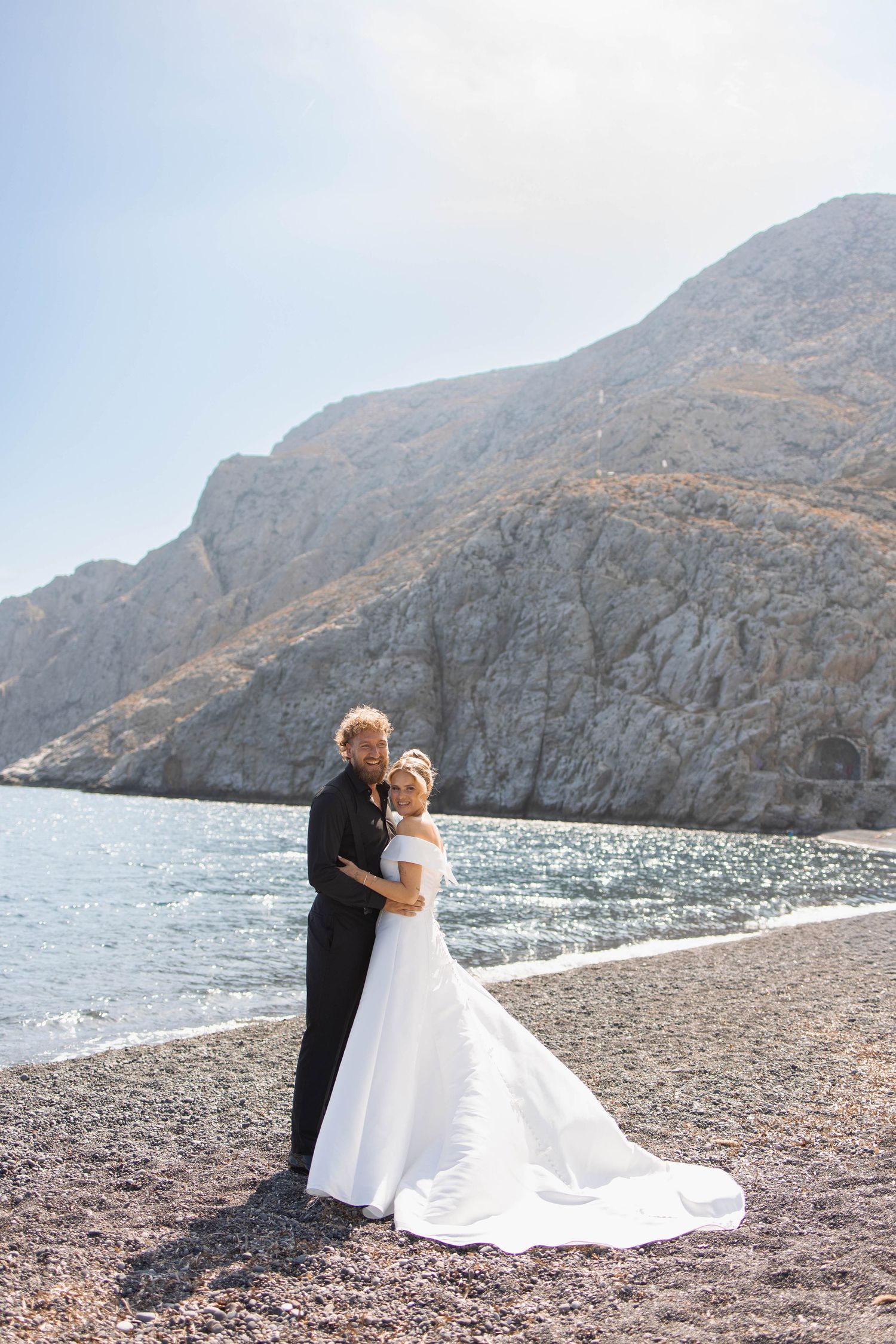 Bride in a white strapless gown embracing a groom in black on a pebble beach with calm sea and rugged cliffs behind them.