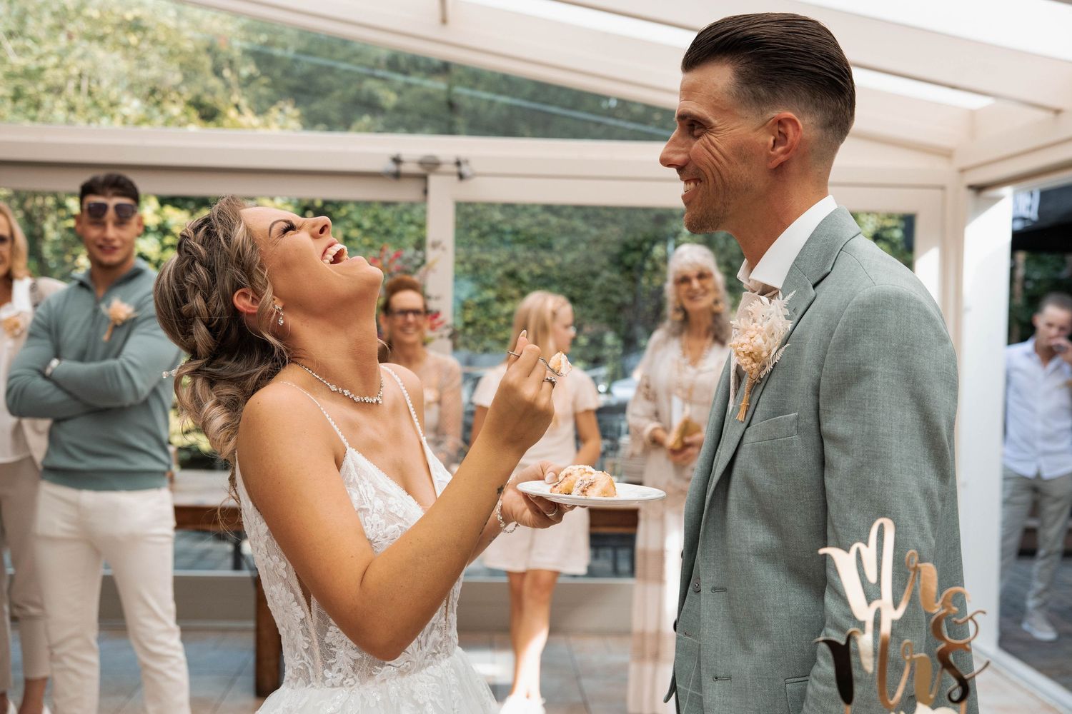Bride and groom laughing joyfully during wedding cake cutting ceremony, guests celebrating in background.