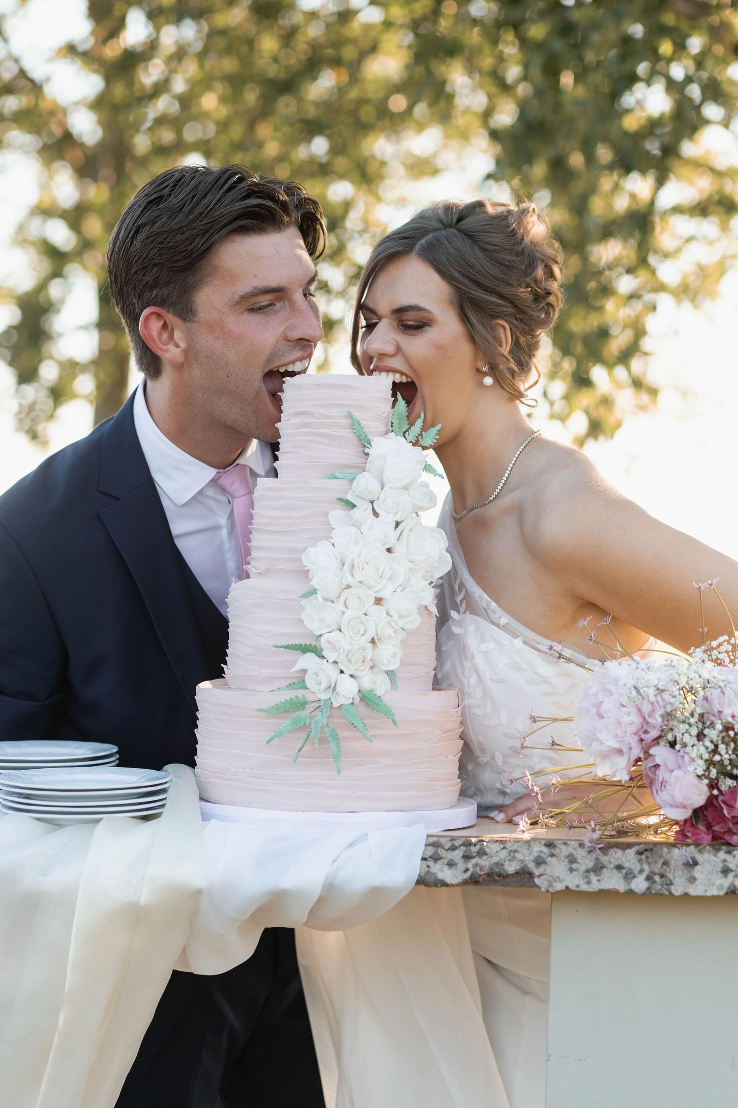 Bride and groom joyfully biting into a elegant pink tiered wedding cake decorated with white flowers outdoors.
