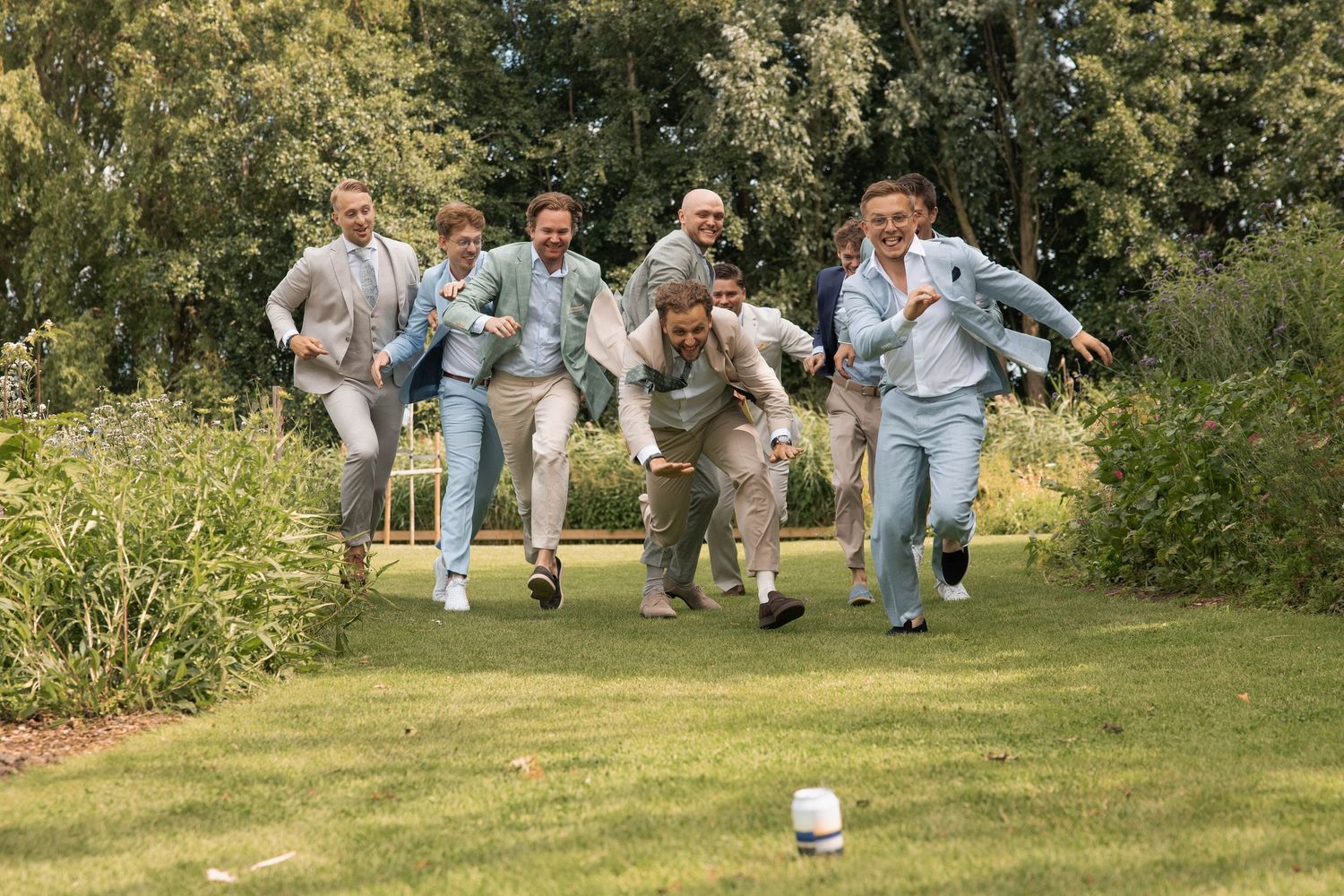 Groomsmen in light blue suits racing across a green lawn at a wedding celebration outdoors.