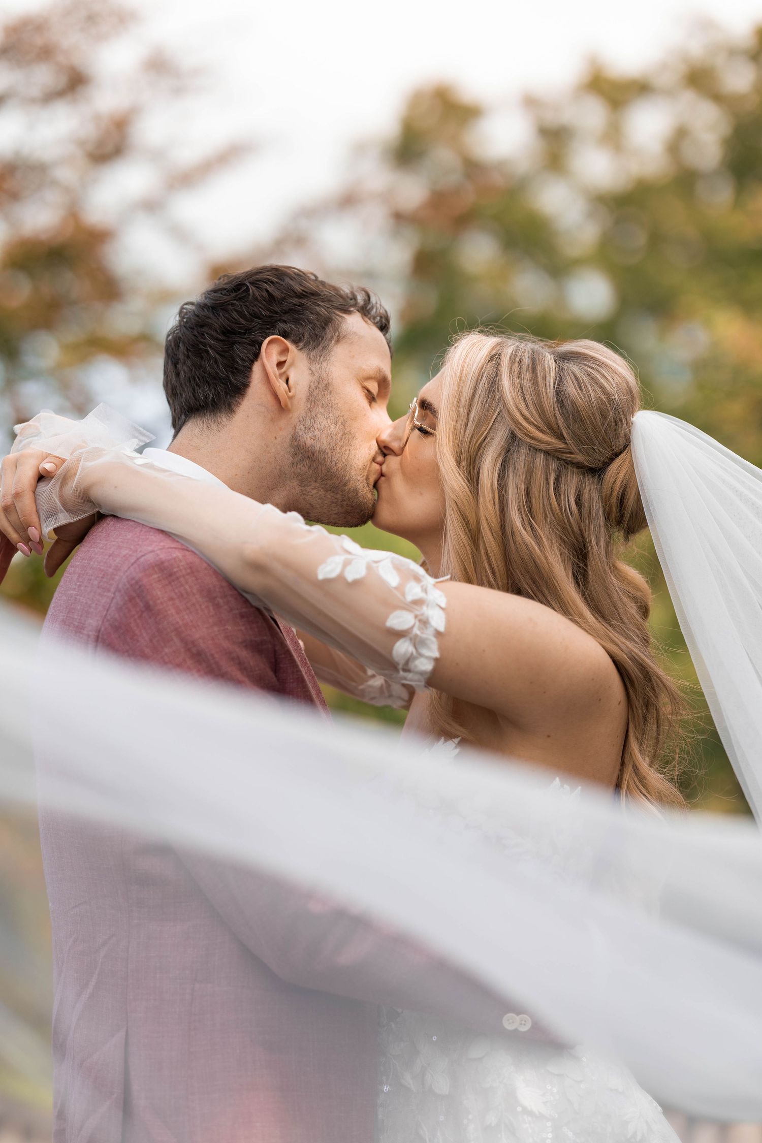 Bride and groom share a romantic kiss outdoors as her sheer lace-trimmed veil billows in the wind against a soft autumn backdrop.