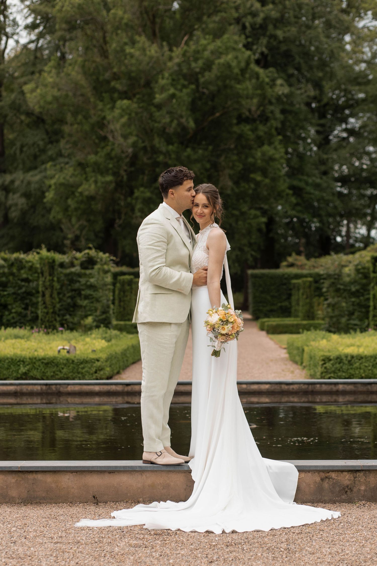 Bride and groom pose elegantly in a formal garden, groom kissing bride's cheek, bride holding bouquet.