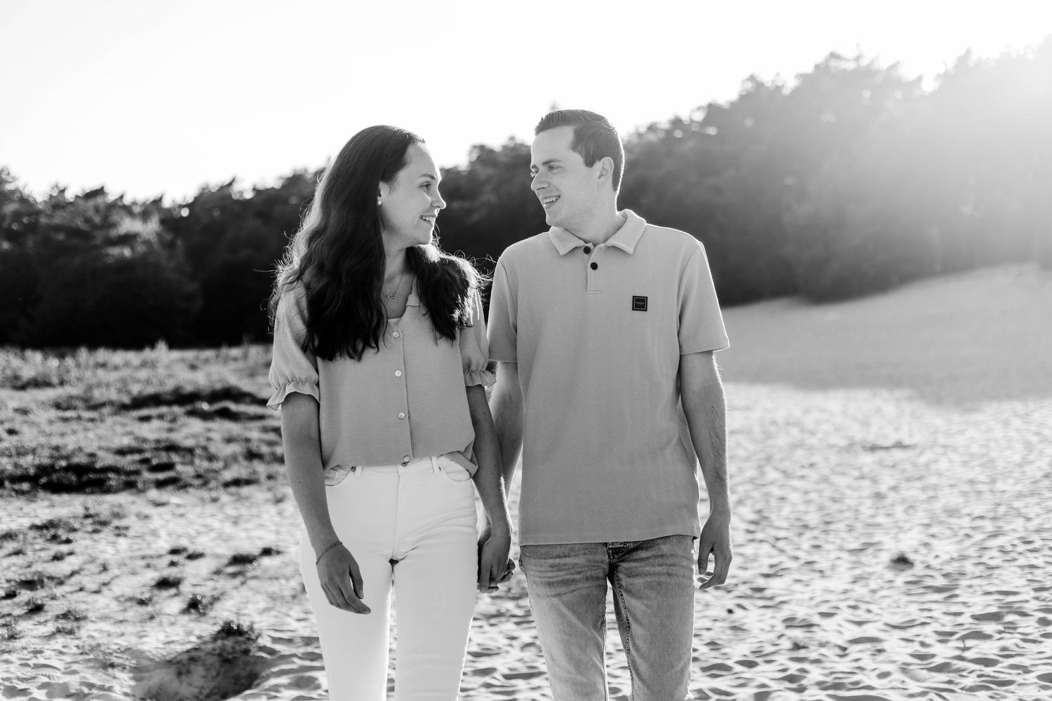 Black and white photo of a couple smiling at each other on a sandy beach with trees in the background.