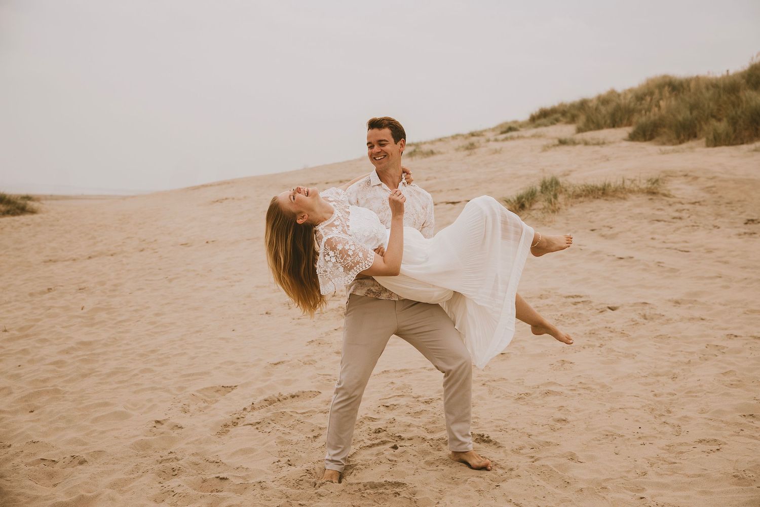 A couple in white outfits dances together on a sandy beach at sunset, creating romantic silhouettes against the sky.