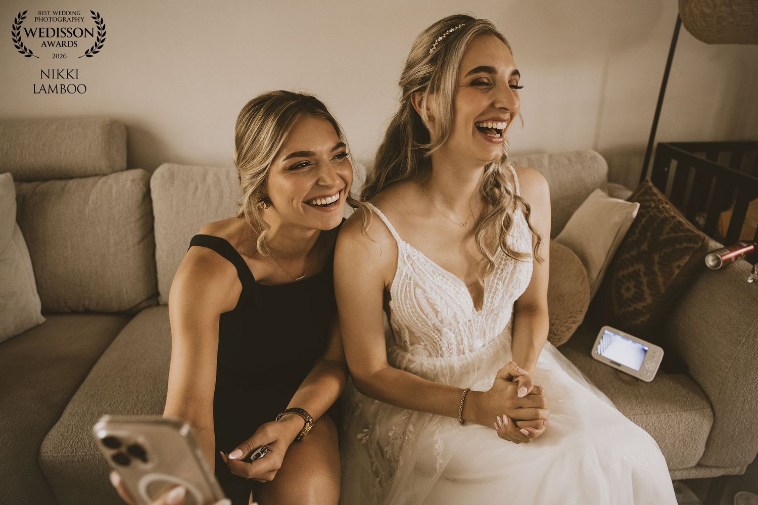 Bride and bridesmaid laughing together on a couch during wedding preparations, captured by Nikko Lambico.
