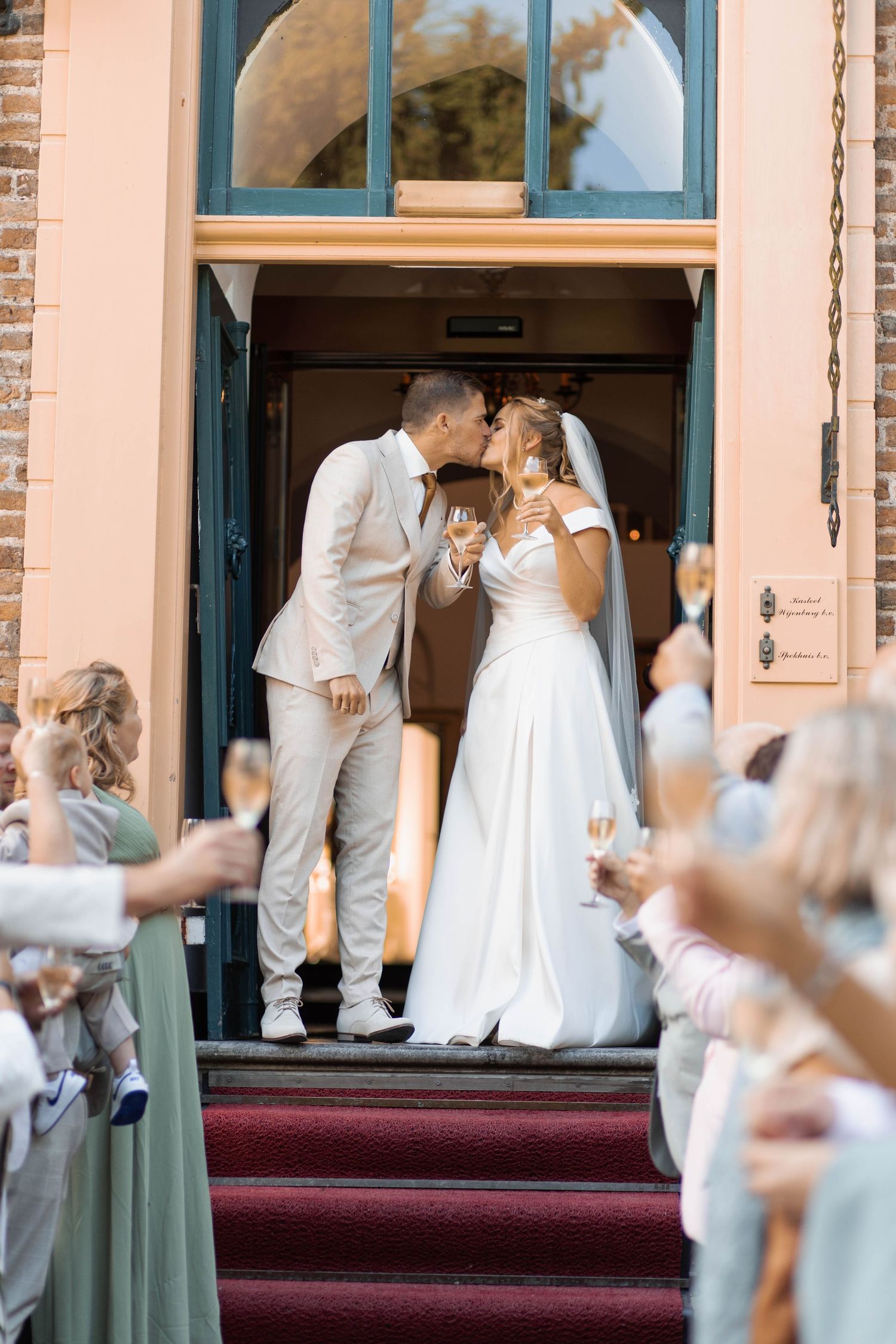 Bride and groom share a kiss on church steps while guests celebrate with champagne glasses at outdoor wedding ceremony.
