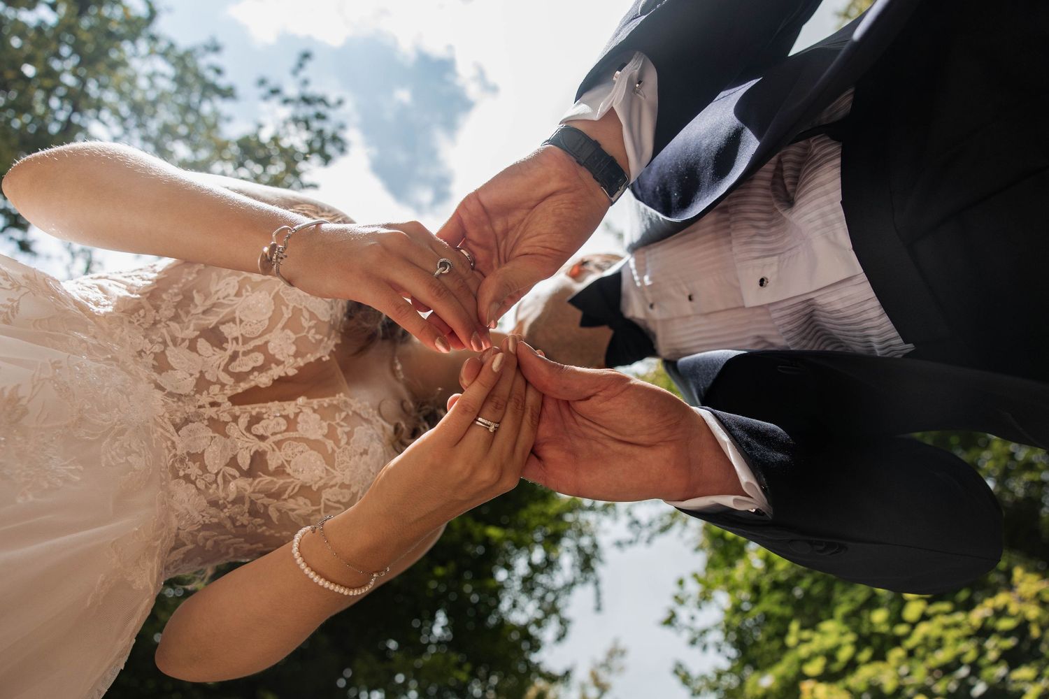 Bride and groom exchanging wedding rings outdoors, close-up of hands with lace dress and formal suit.