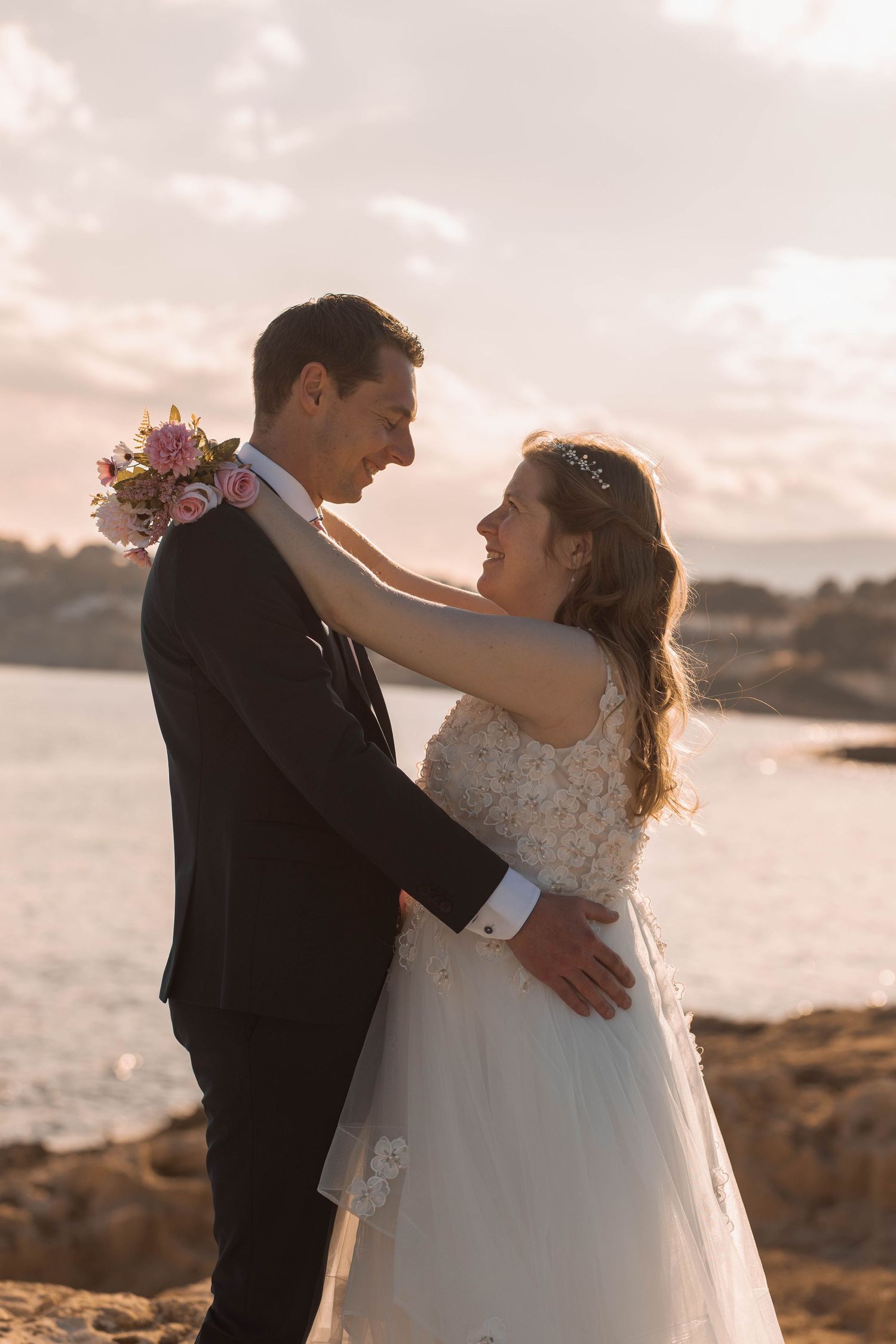 Bride and groom share a romantic moment at sunset by the sea, with the bride holding pink roses.