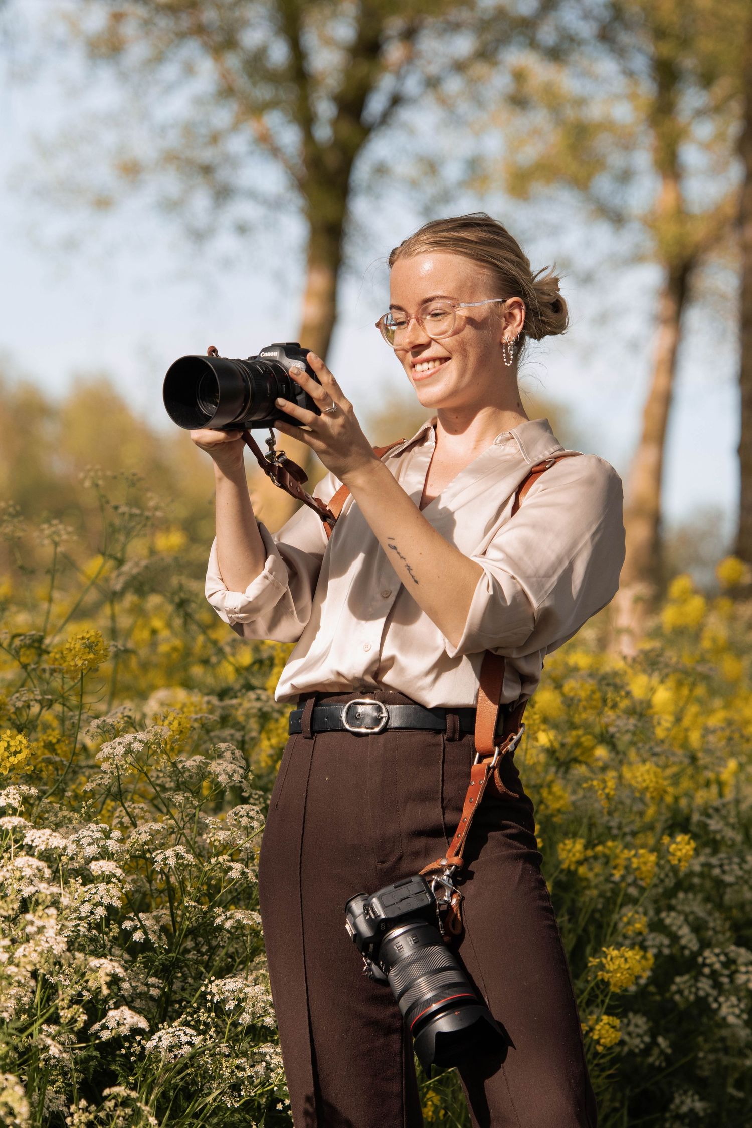 Smiling female photographer reviewing photos on DSLR camera outdoors in blooming wildflower field on sunny day.