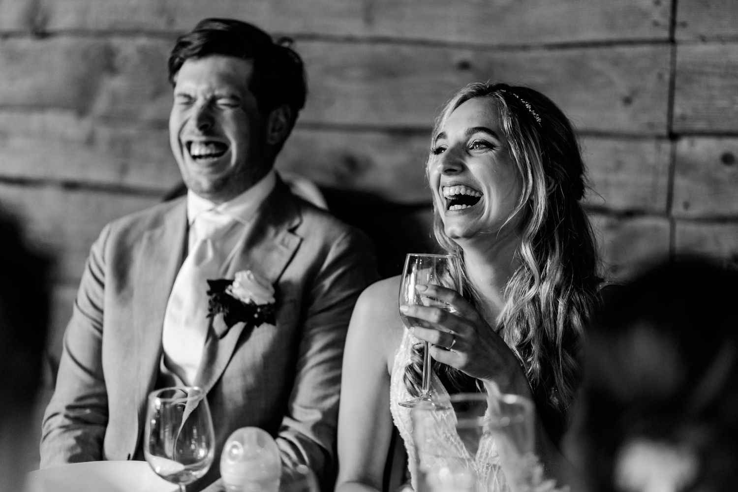 Black and white photo of a laughing bride and groom holding champagne glasses at their wedding reception.