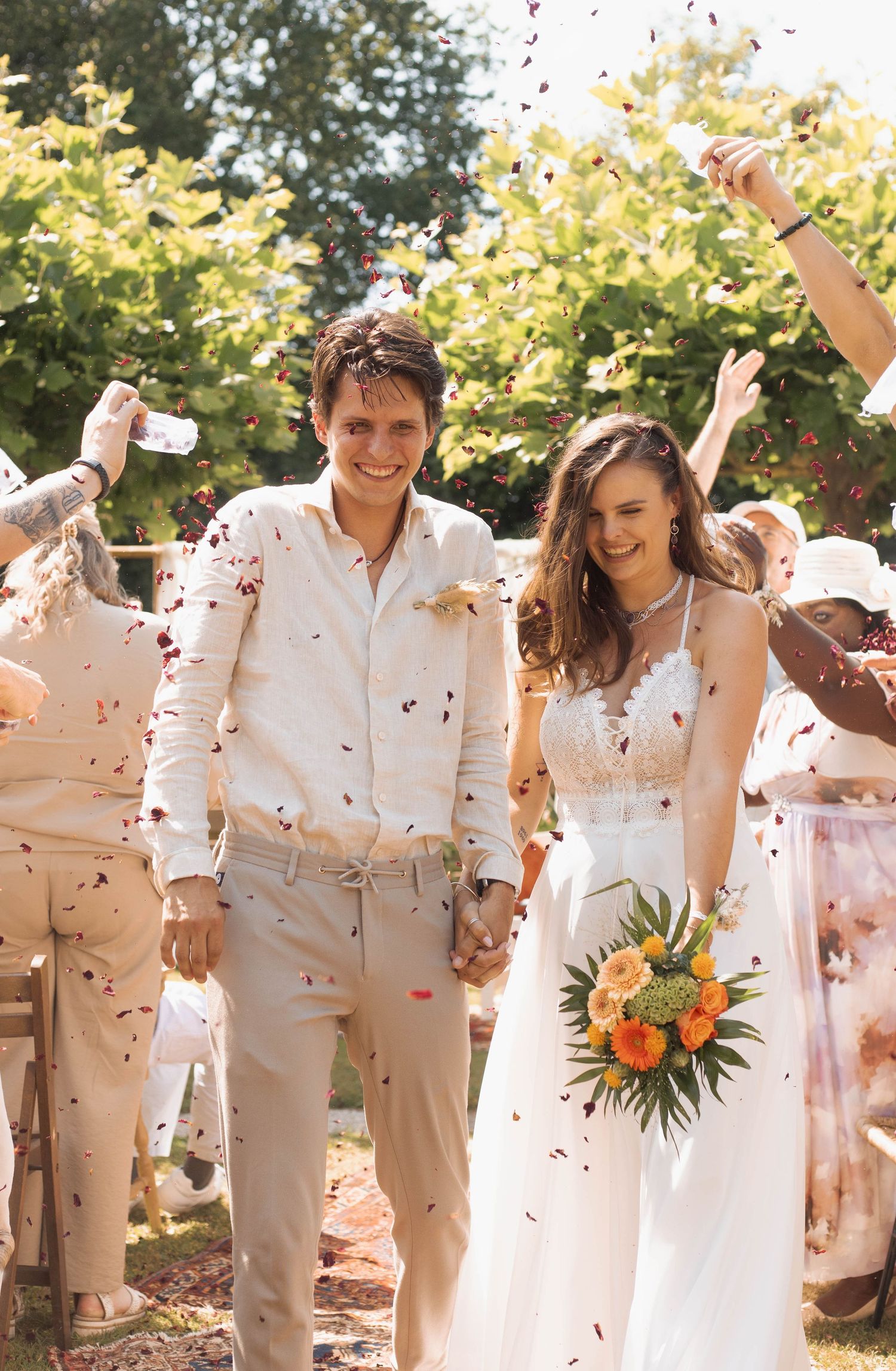 Joyful newlyweds walking through confetti shower, bride holding orange bouquet in white gown, groom in beige suit.
