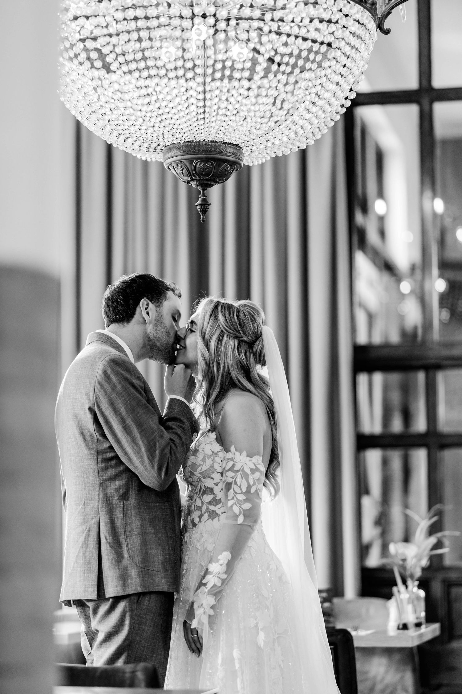 Black and white photo of a bride and groom sharing a kiss beneath an elegant crystal chandelier in a romantic venue.