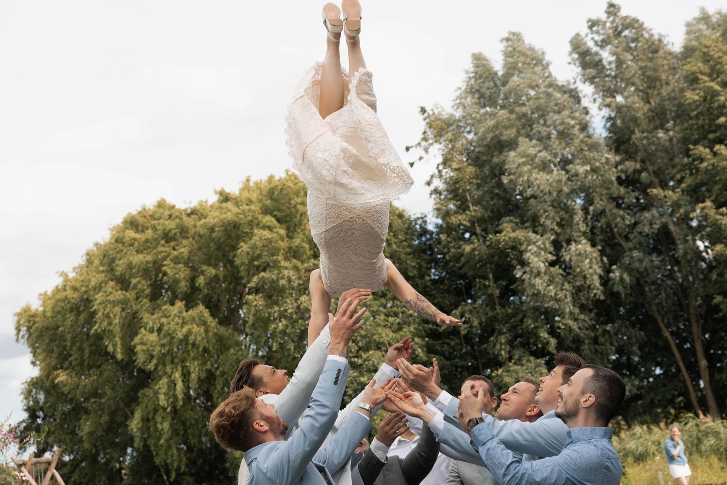 Groomsmen in light blue suits tossing bride in wedding dress into the air outdoors.