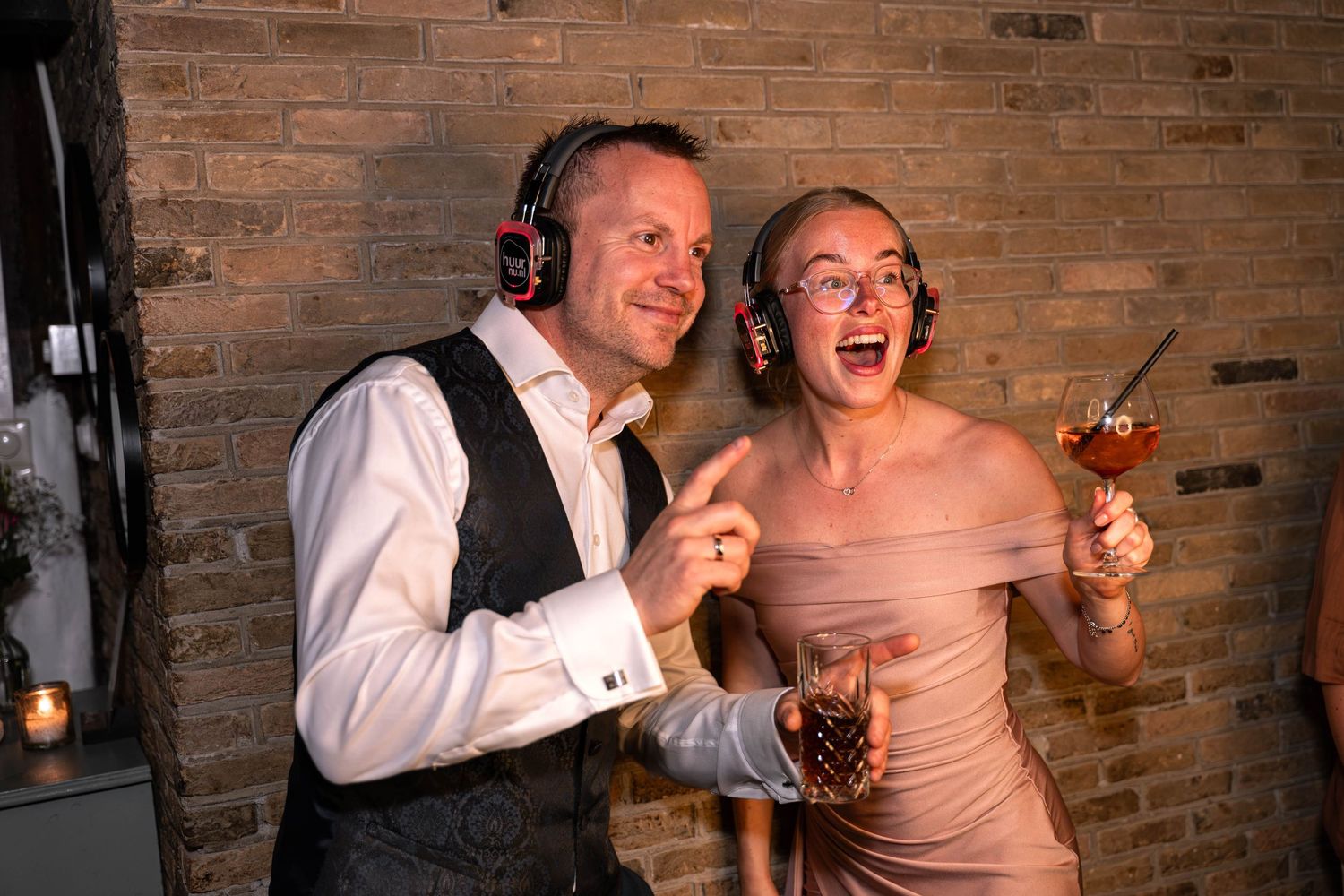 Couple wearing headphones laughing and holding cocktails at a party near a brick wall.
