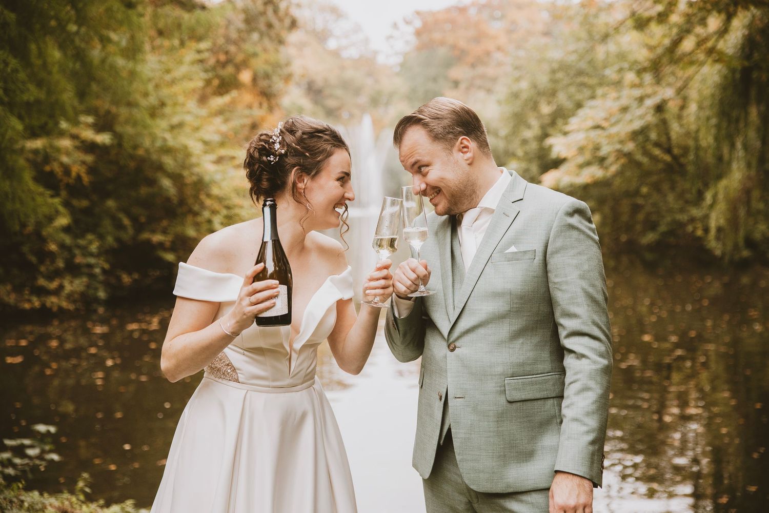 A couple shares a laugh while holding champagne glasses in an outdoor garden setting with warm autumn colors.