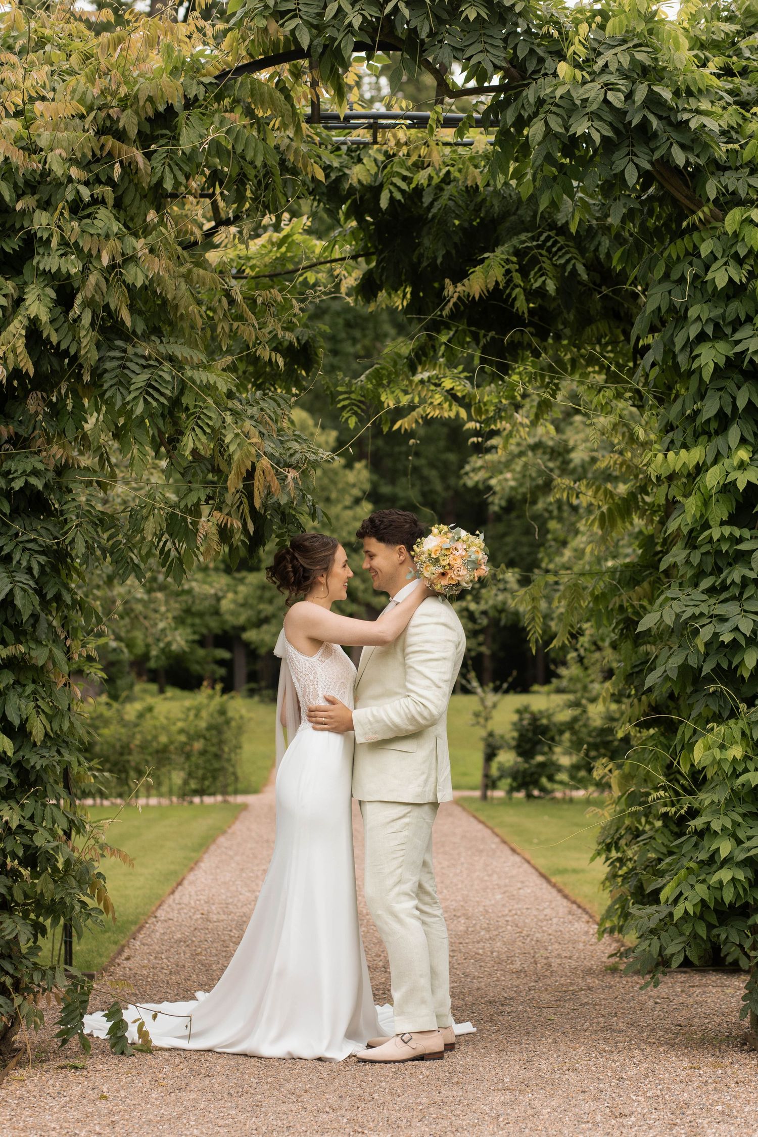Bride and groom embrace under a lush green garden arch on a gravel path on their wedding day.