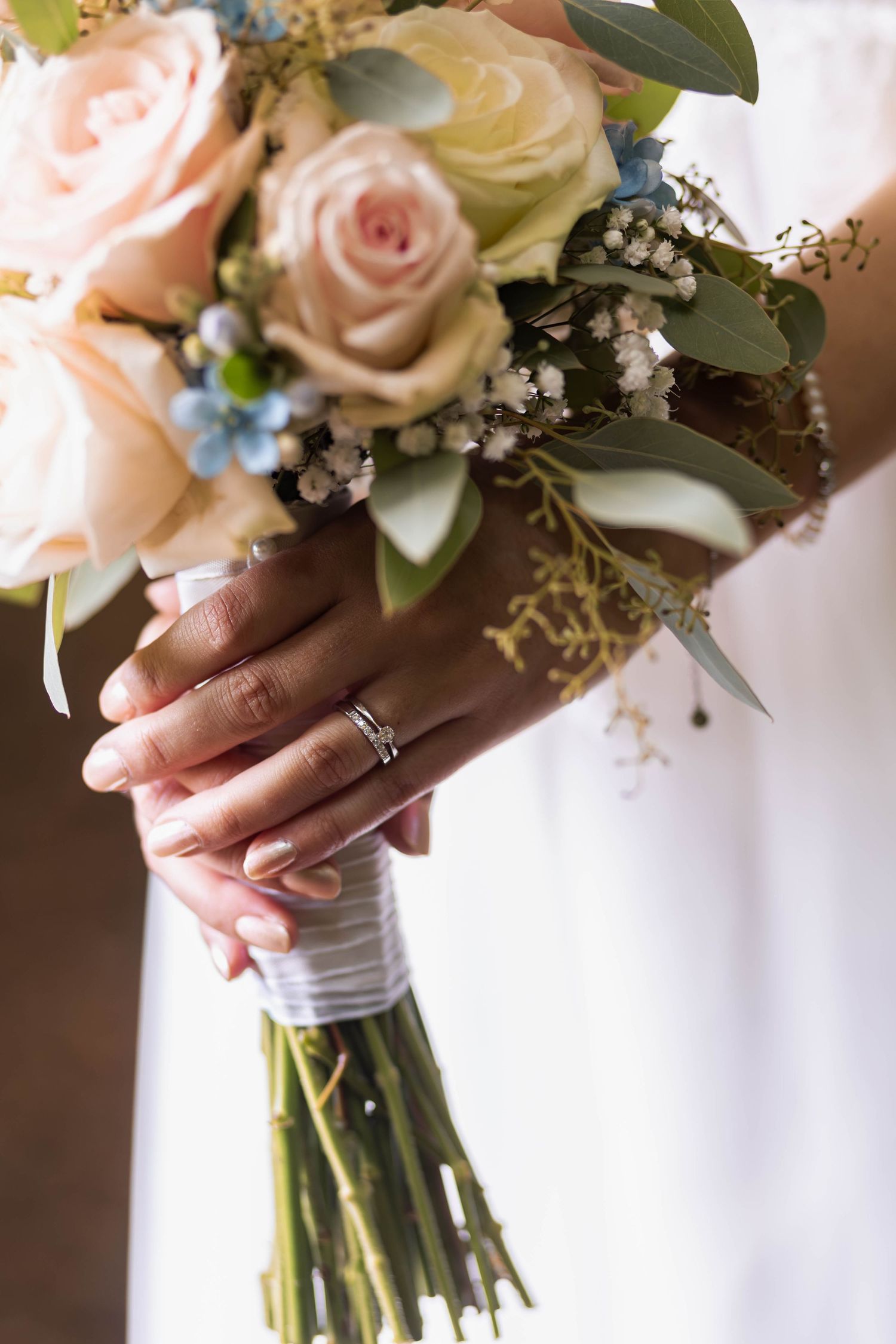 Bride holding a pastel wedding bouquet with pink roses and blue berries, wearing a diamond wedding ring.
