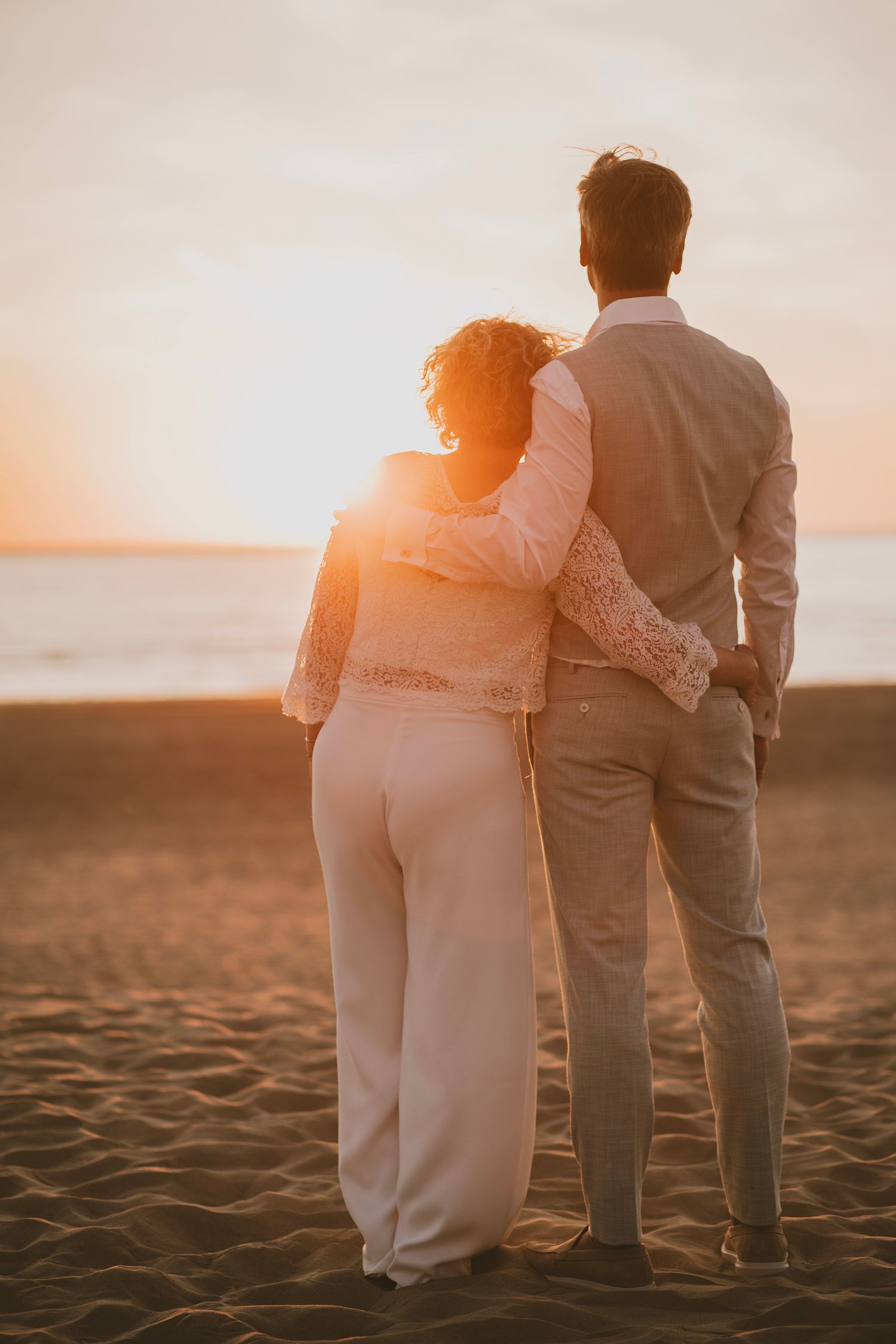 A couple stands together on a sandy beach at sunset, silhouetted against the warm golden light.