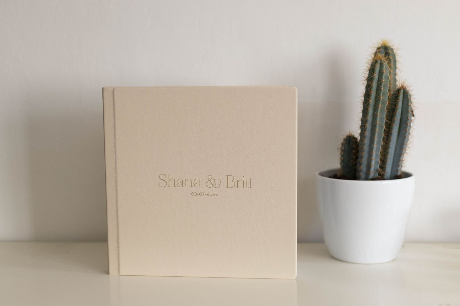 Personalized beige wedding photo album for Shane and Bria displayed on a white shelf next to a small cactus plant.