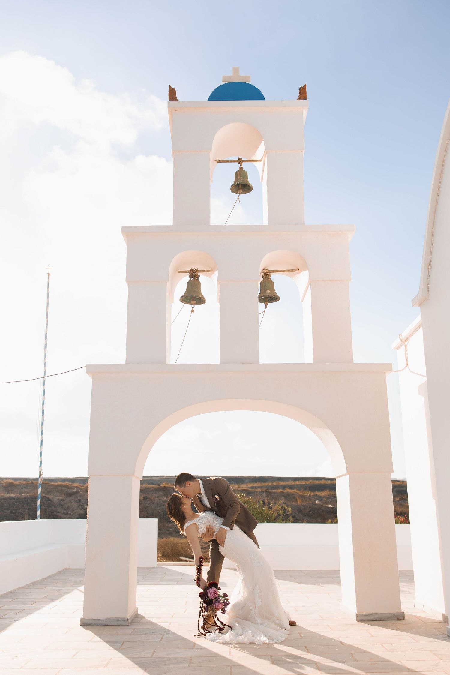 Bride and groom share a romantic dip kiss beneath a white arched bell tower with a blue dome on a sunny Greek island.