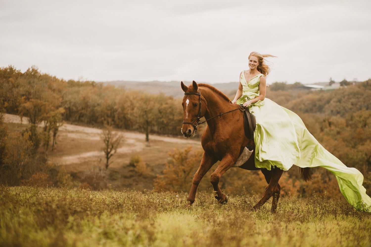 A person in a flowing yellow dress sits atop a brown horse in a scenic autumn meadow landscape at sunset.