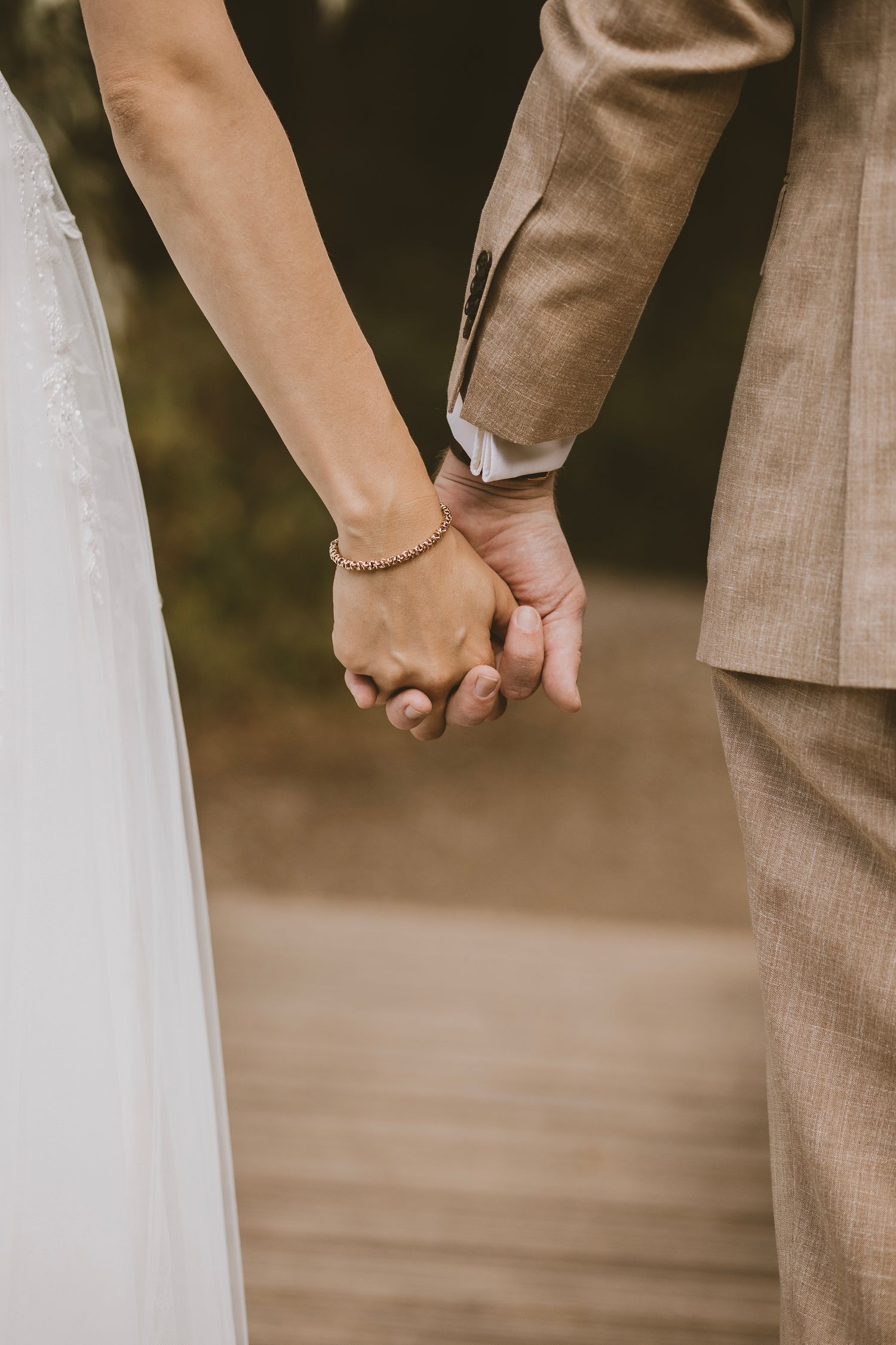 Close-up of newlyweds holding hands during wedding ceremony, with bride in white dress and groom in beige suit.