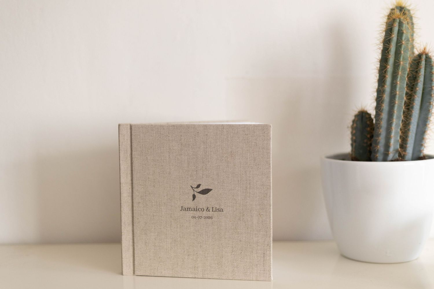 Linen-covered wedding photo album with floral monogram sits on a white shelf beside a tall cactus in a white pot.