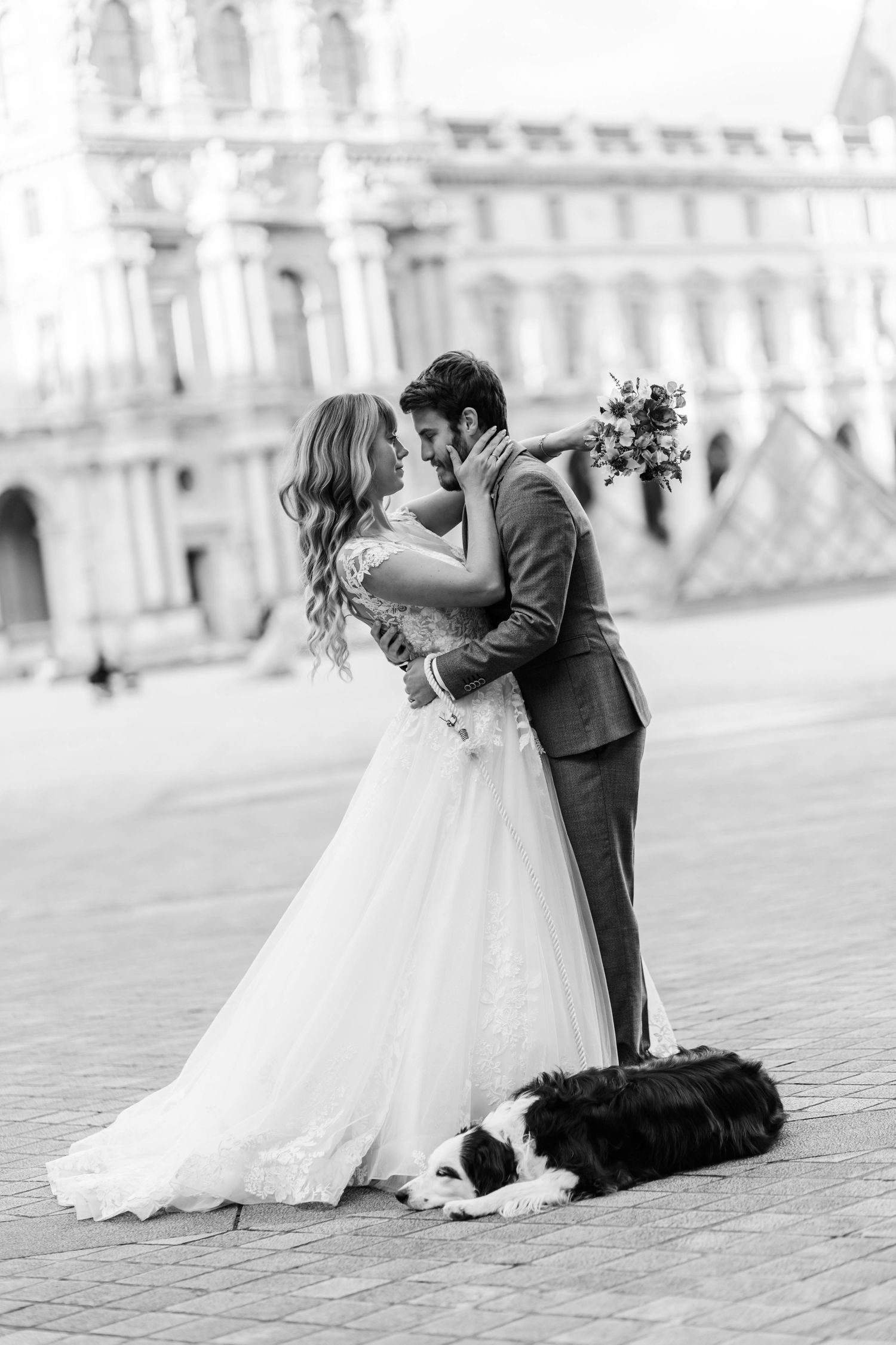 Black and white photo of a bride and groom kissing in front of the Louvre in Paris, with a dog resting at their feet.