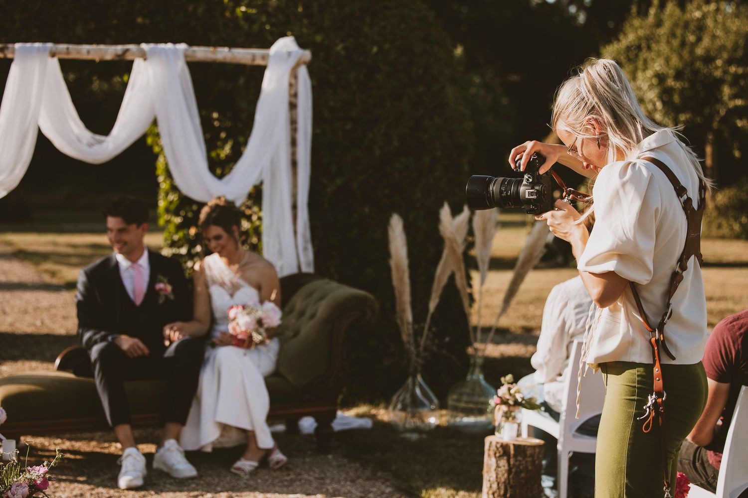 A photographer captures a romantic outdoor wedding ceremony under a white draped arch in warm sunlight.