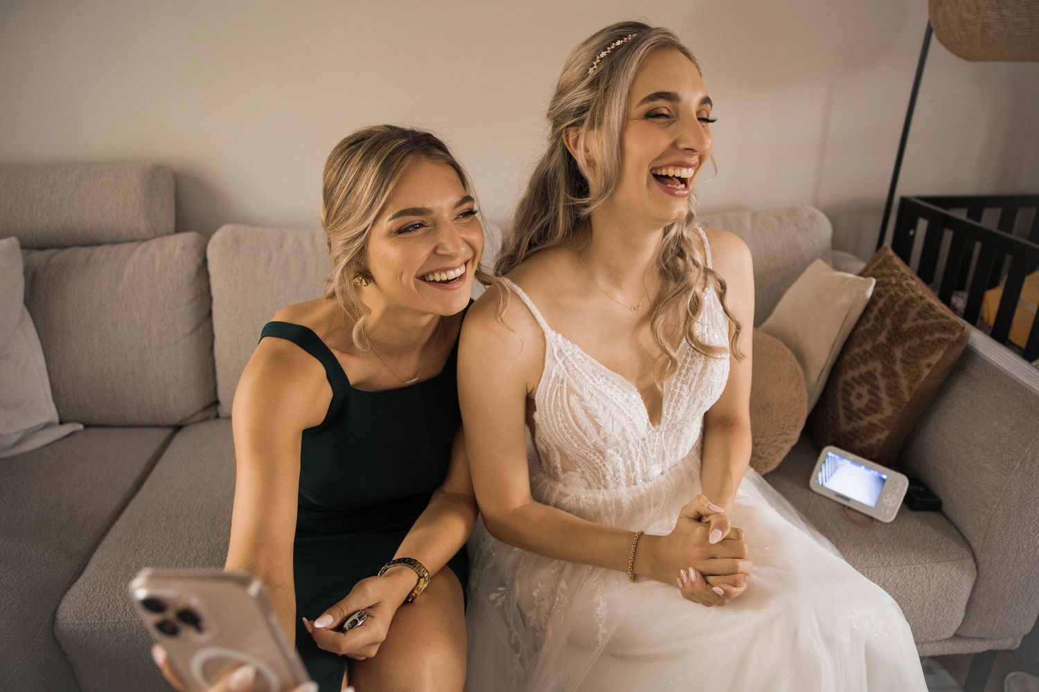 Two laughing women sit on a gray couch, one in a white wedding dress and one in a black dress, taking a selfie.