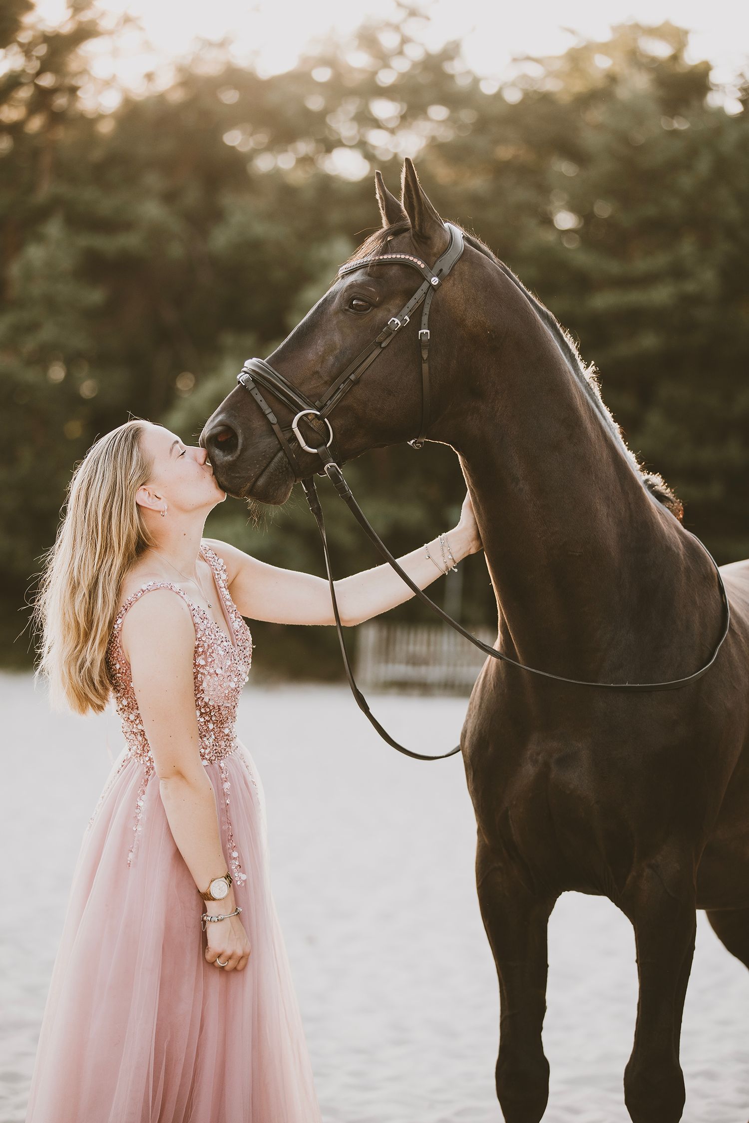 A person in a pink flowy dress shares a tender moment with a black horse during a golden hour photoshoot.