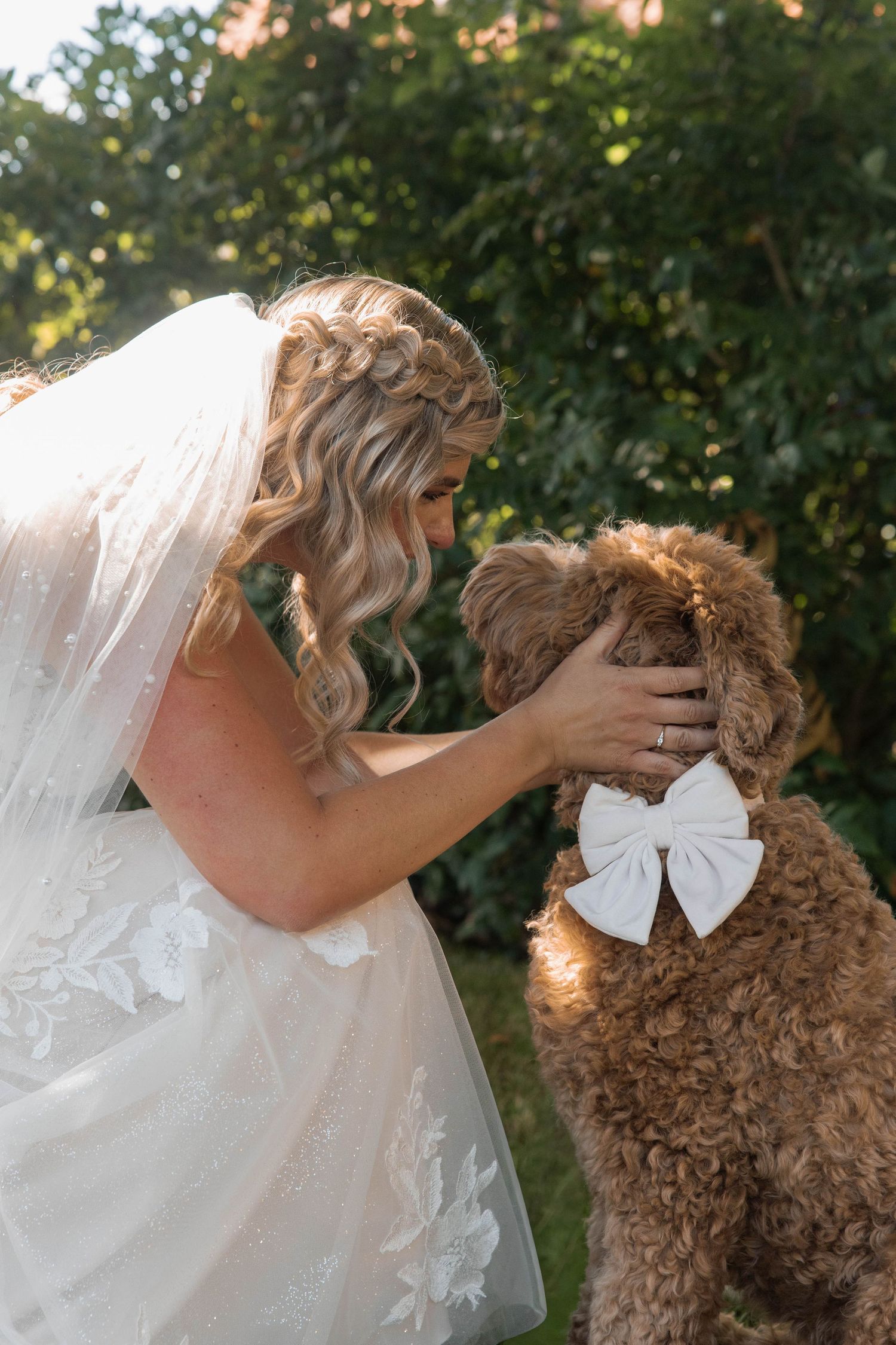 Bride in white lace gown and veil tenderly touches a curly brown doodle dog wearing a white bow outdoors.