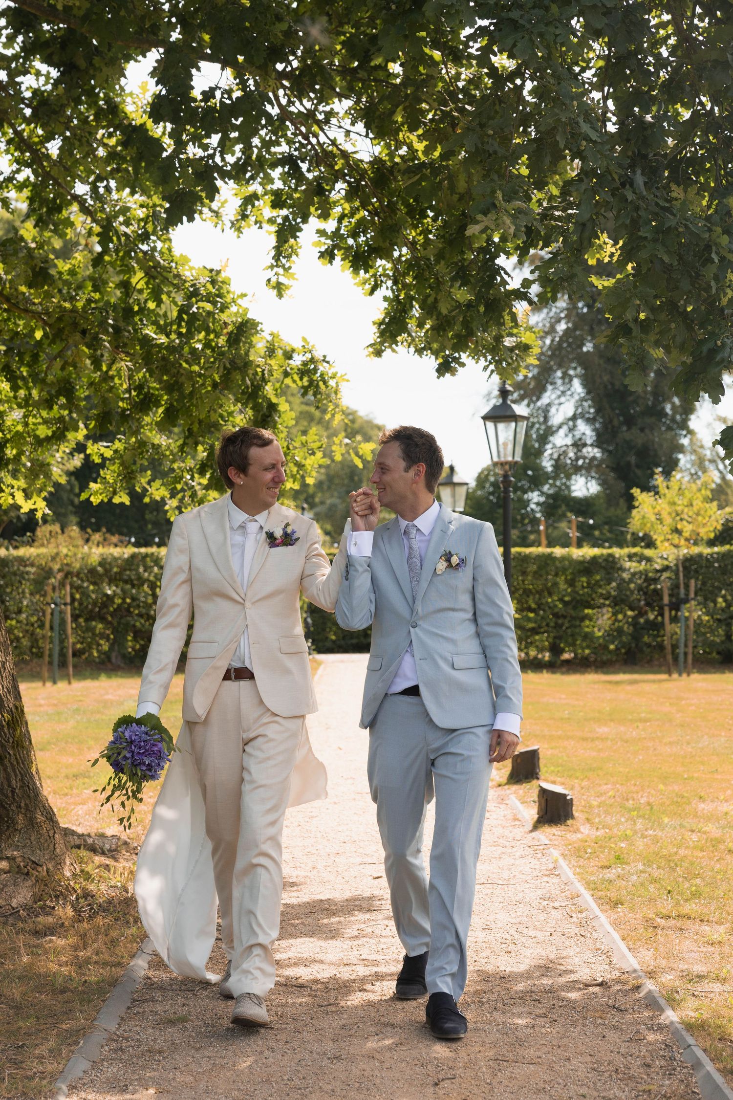 Two grooms in suits walk hand in hand along a sunny garden path, smiling at each other on their wedding day.