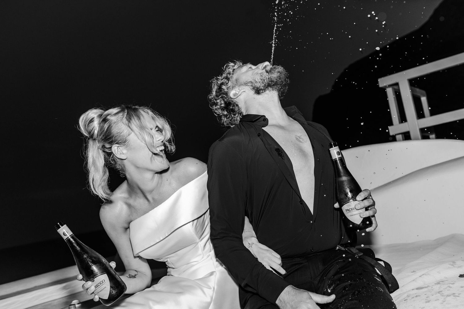 Black and white photo of a laughing bride in a white gown and a groom spraying champagne into his mouth while celebrating at night.