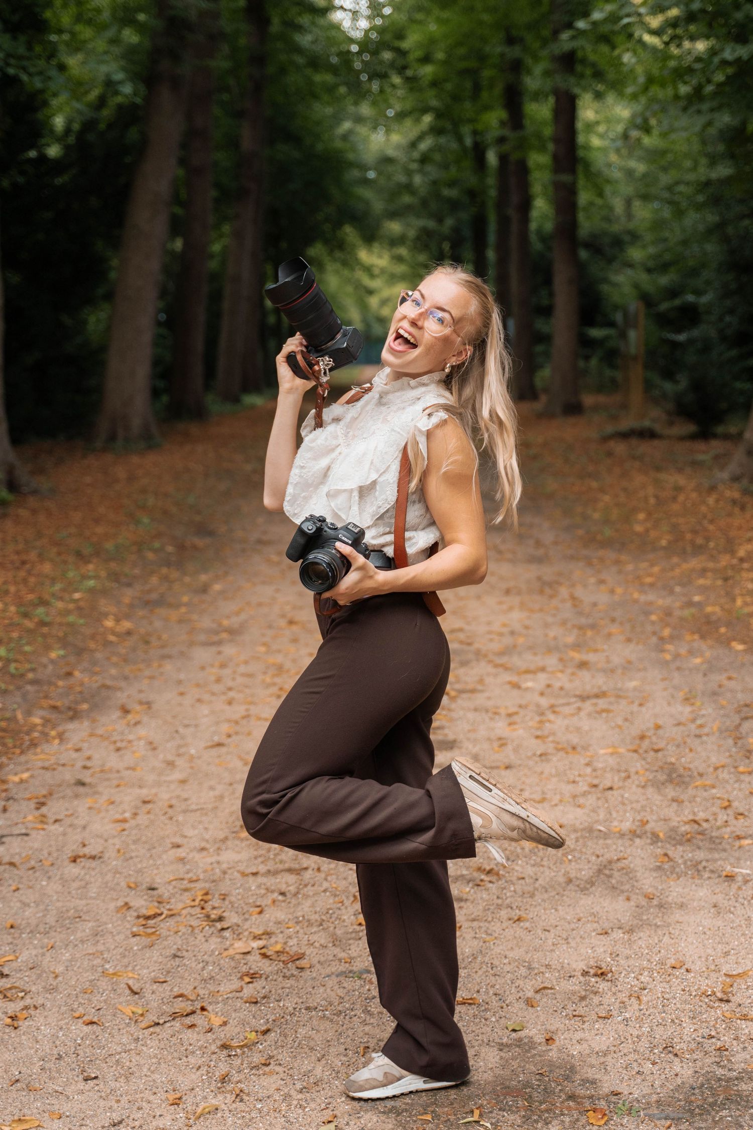 Joyful blonde female photographer holding cameras on a forest path, wearing brown pants and white blouse.