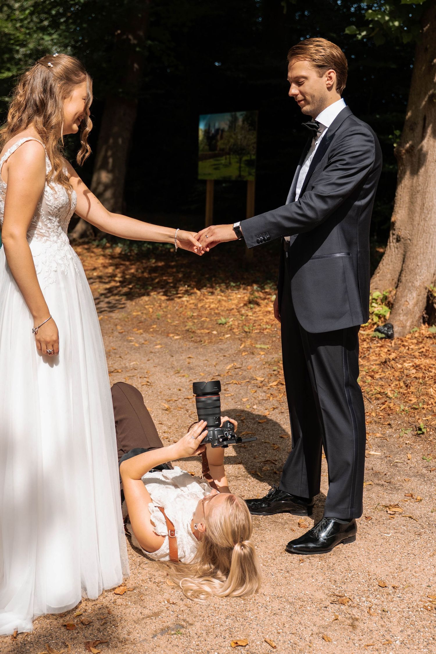 Photographer crouching to capture bride and groom holding hands in a sunny outdoor park setting during wedding shoot.