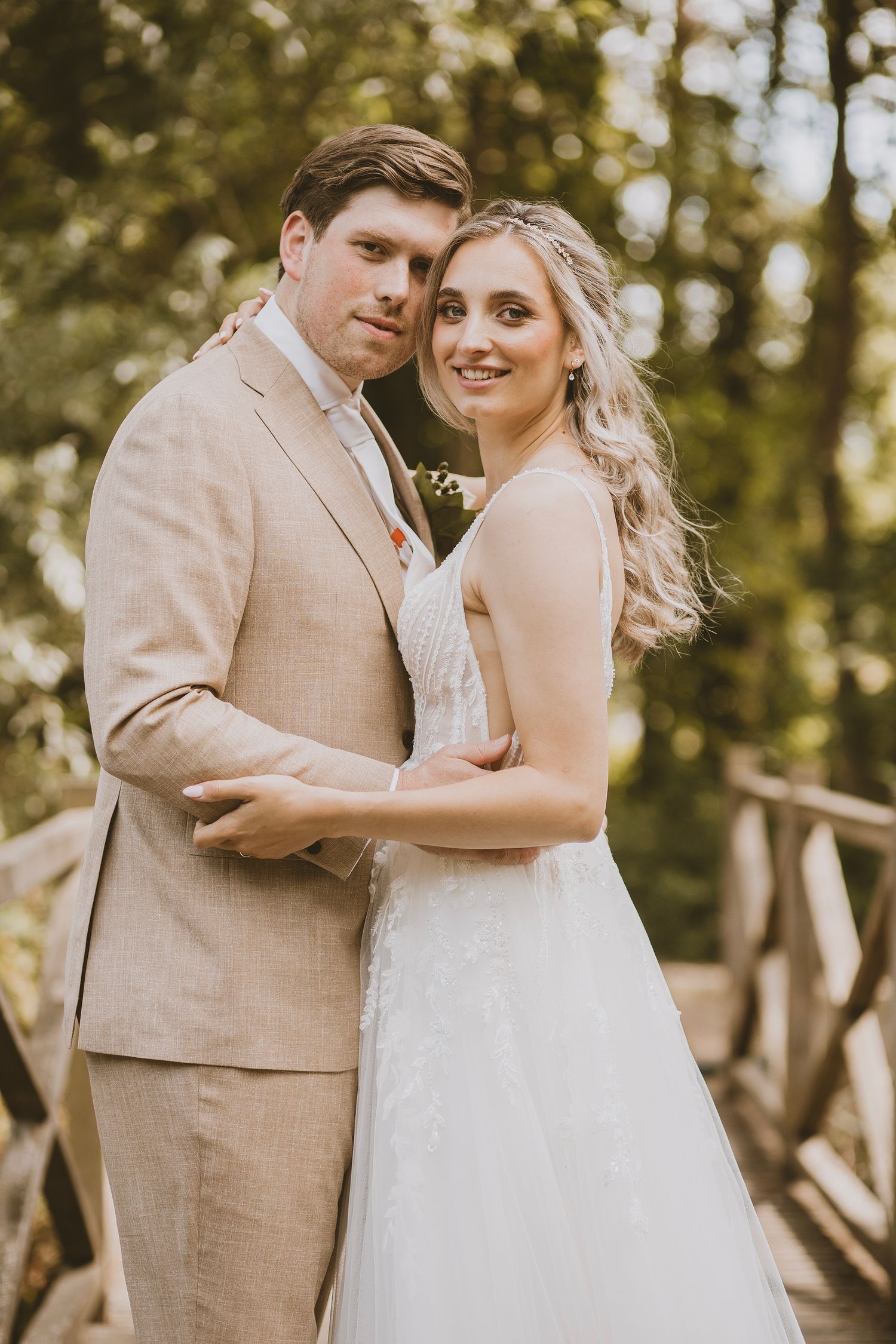 A romantic wedding portrait of a couple in a tan suit and white dress embracing in a sunlit wooded setting.