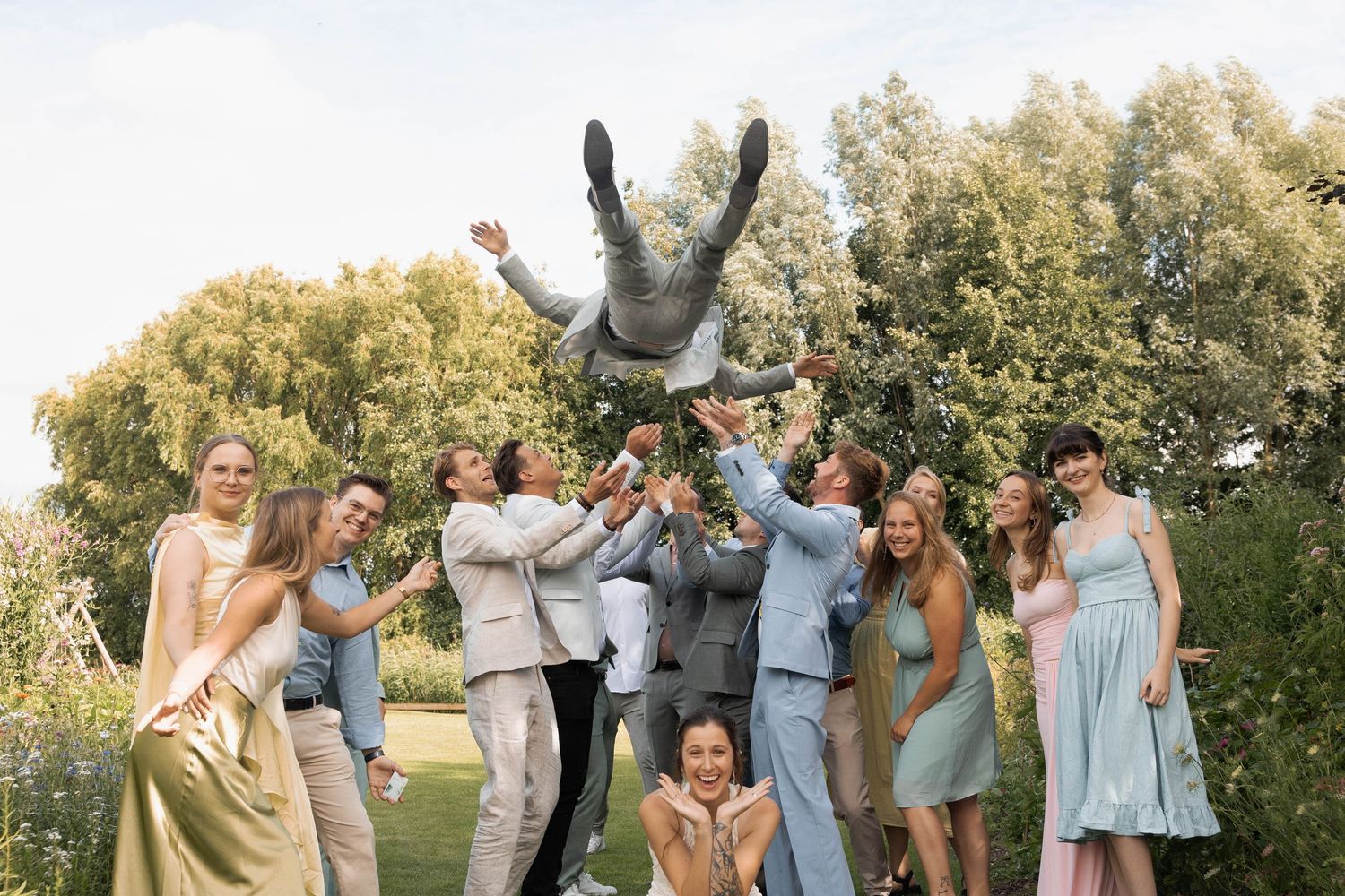Wedding guests tossing groom into air outdoors with trees in background.