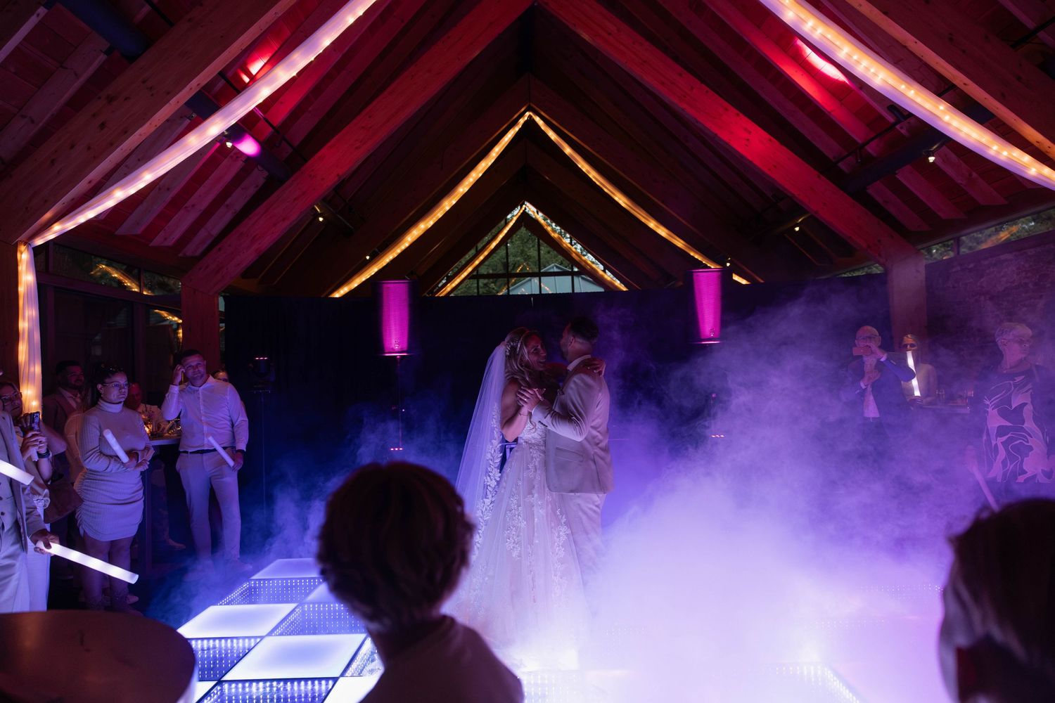 Bride and groom share first dance on illuminated floor in rustic barn venue with purple fog effects.