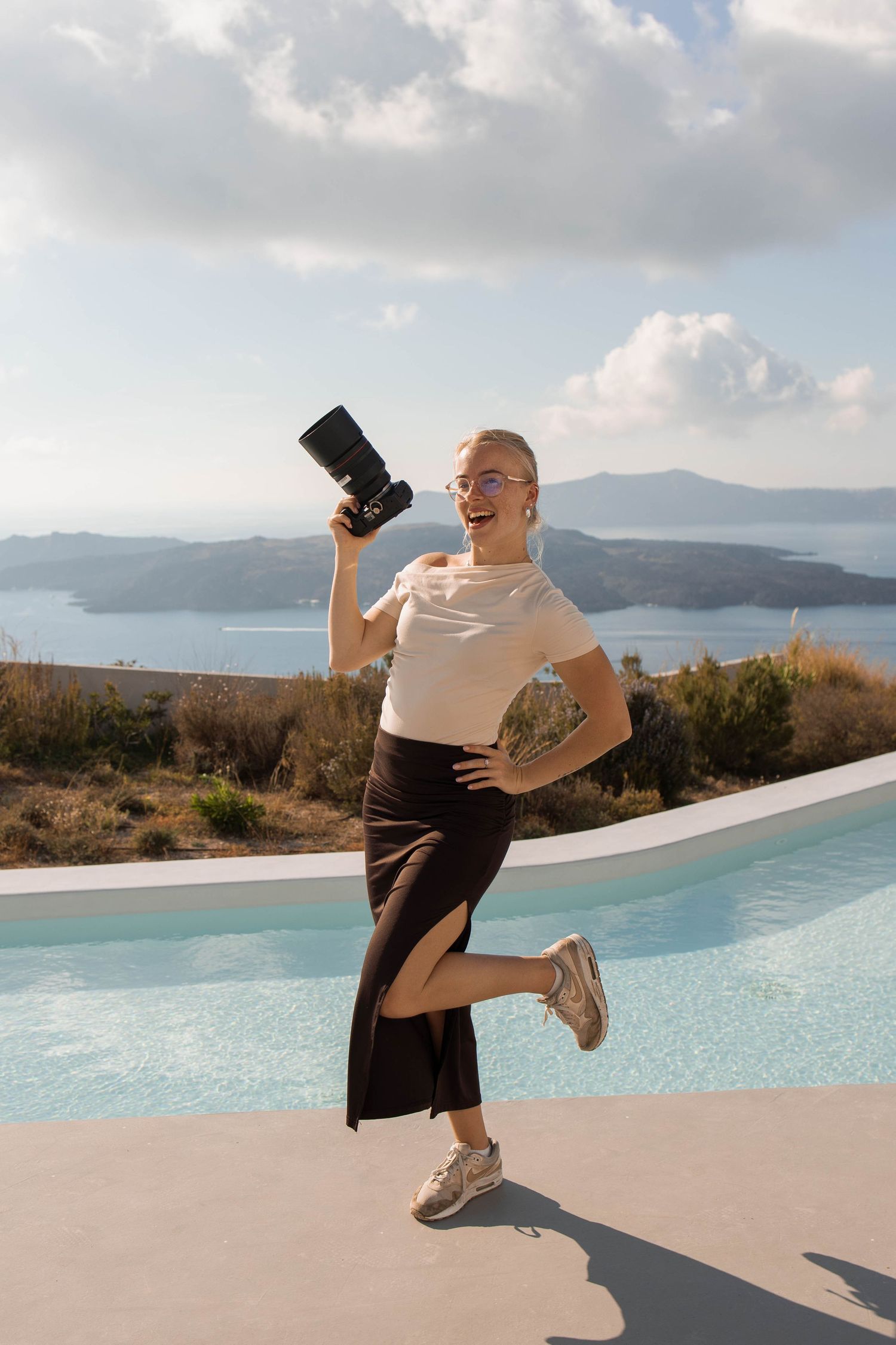 Smiling woman in a white top and black skirt holds a camera and poses playfully beside a pool overlooking the sea and islands.