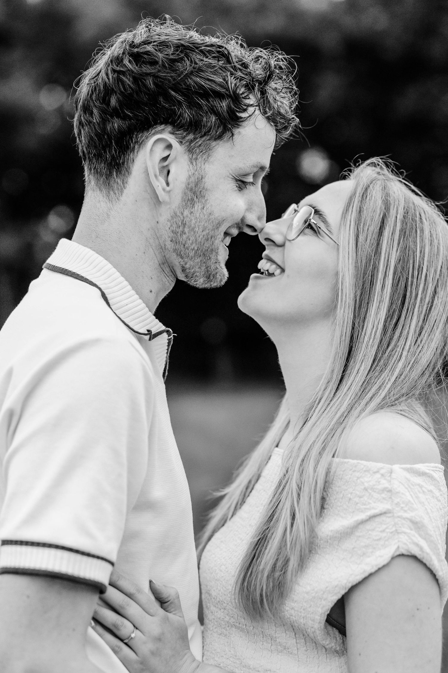 Black and white photo of a smiling couple leaning in for a kiss outdoors in a romantic moment.