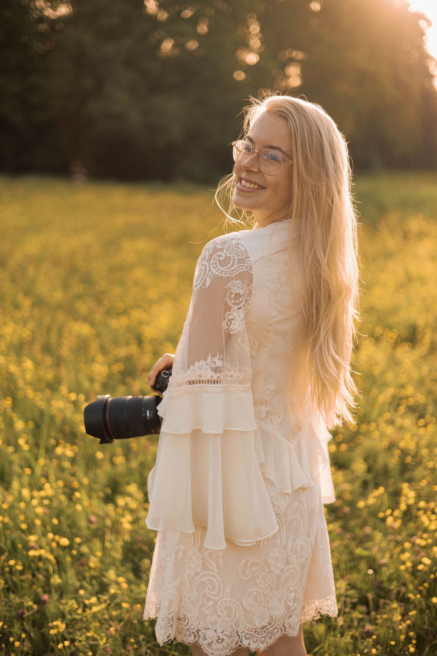 Smiling blonde woman in white lace dress holding camera in golden sunlit flower field at sunset.